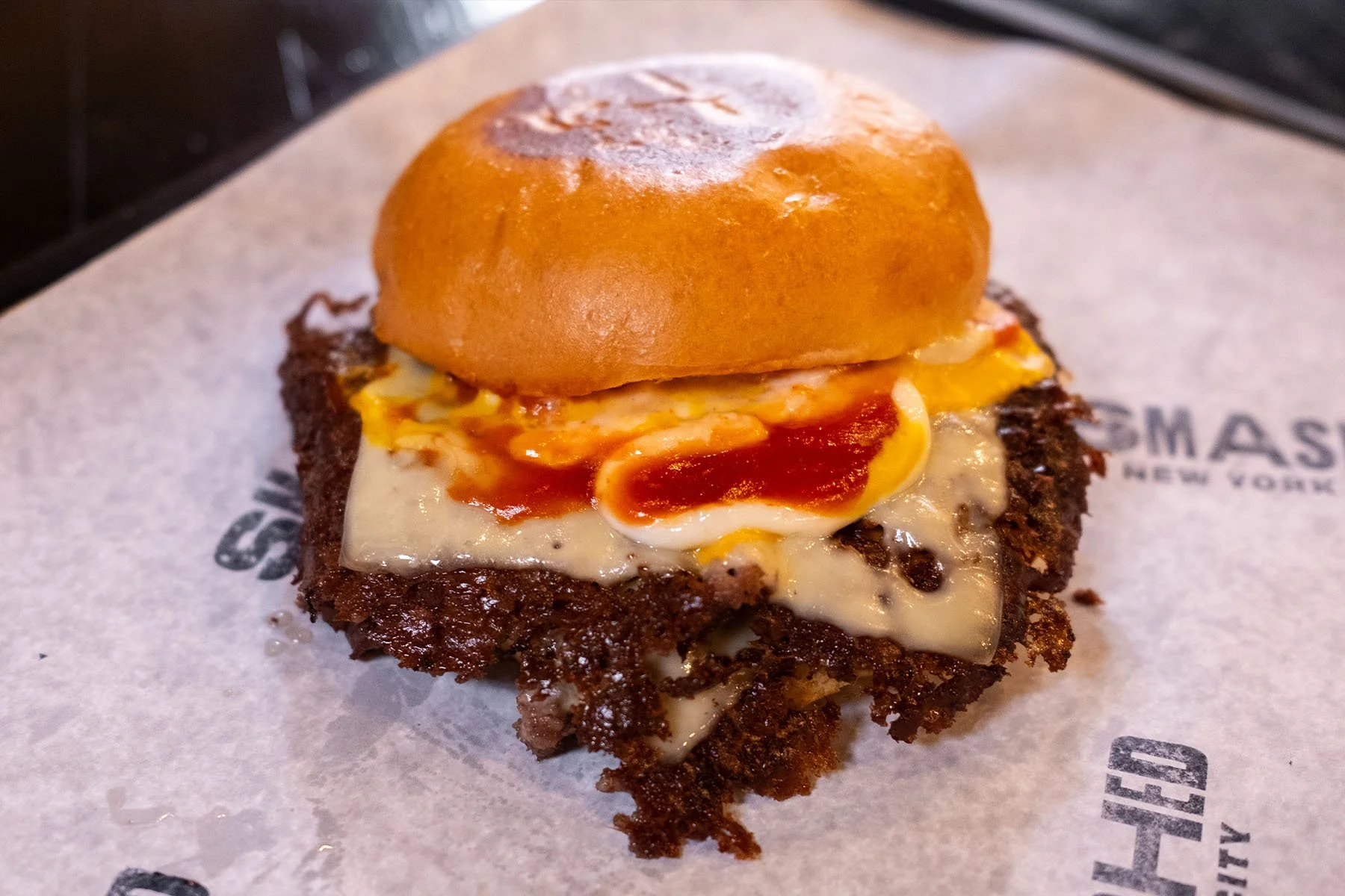A hamburger with a toasted bun, fried egg, melted cheese, spicy sauce, and a beef patty served on branded paper.