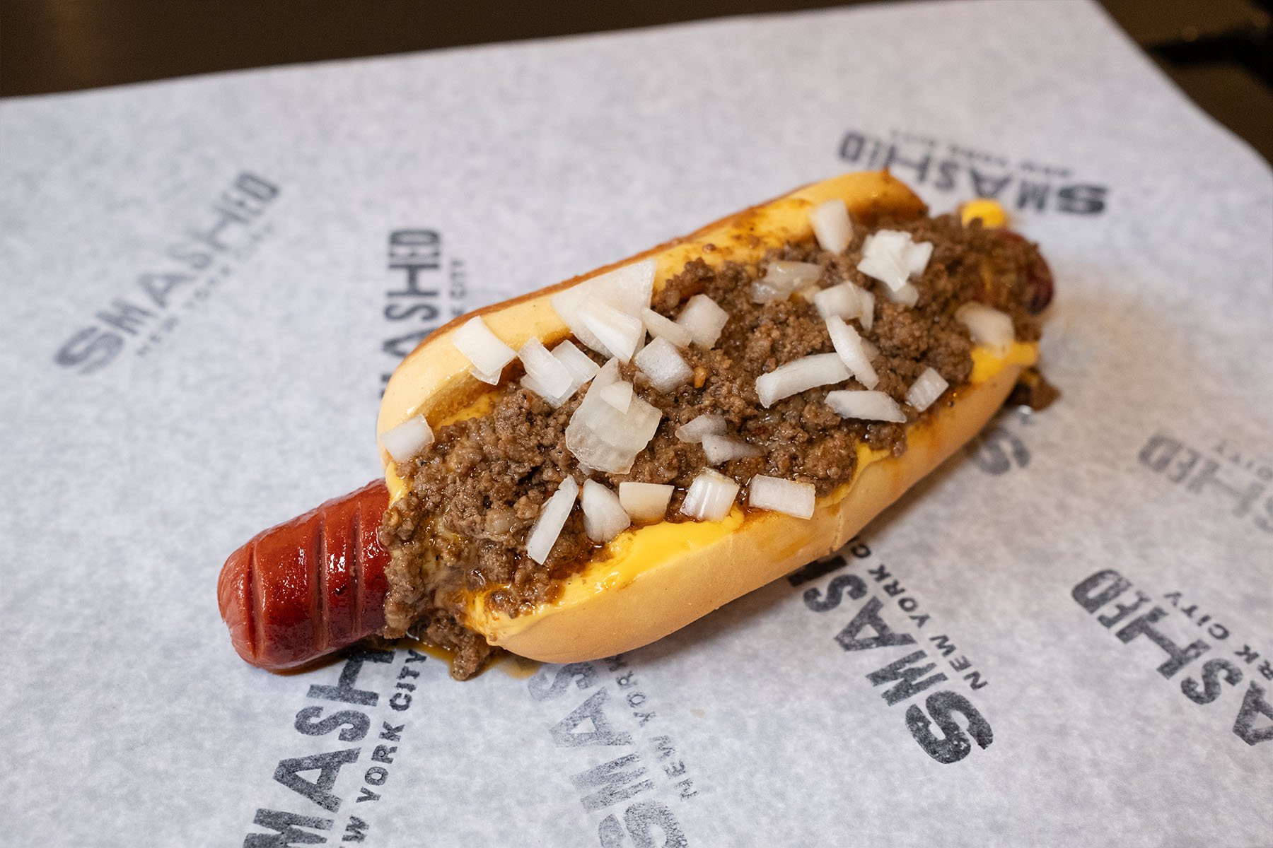Hot dog with diced onions, ground beef, and mustard on a piece of branded paper from Shake Shack.