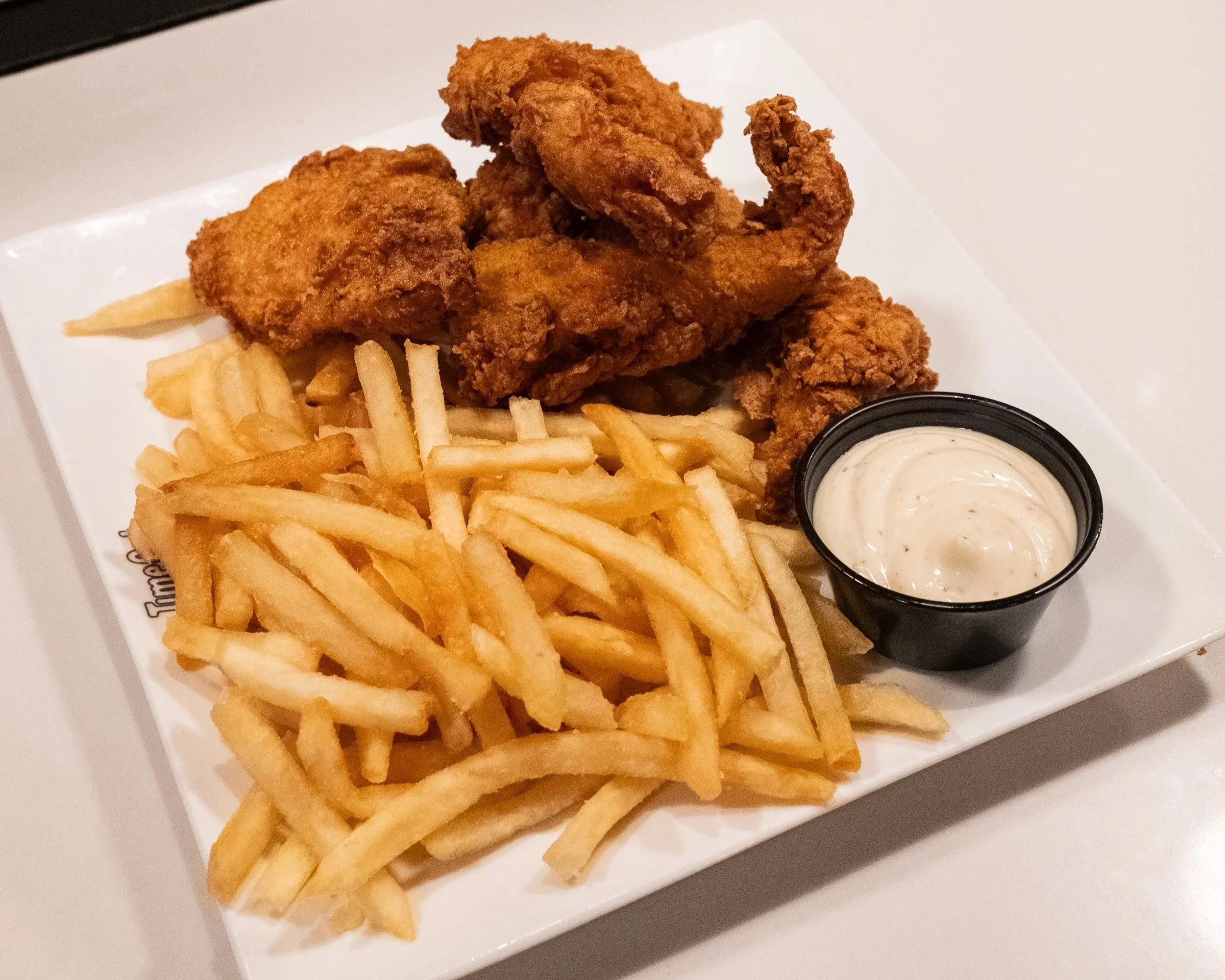 Fried chicken tenders with French fries and a small cup of ranch dressing on a white square plate.