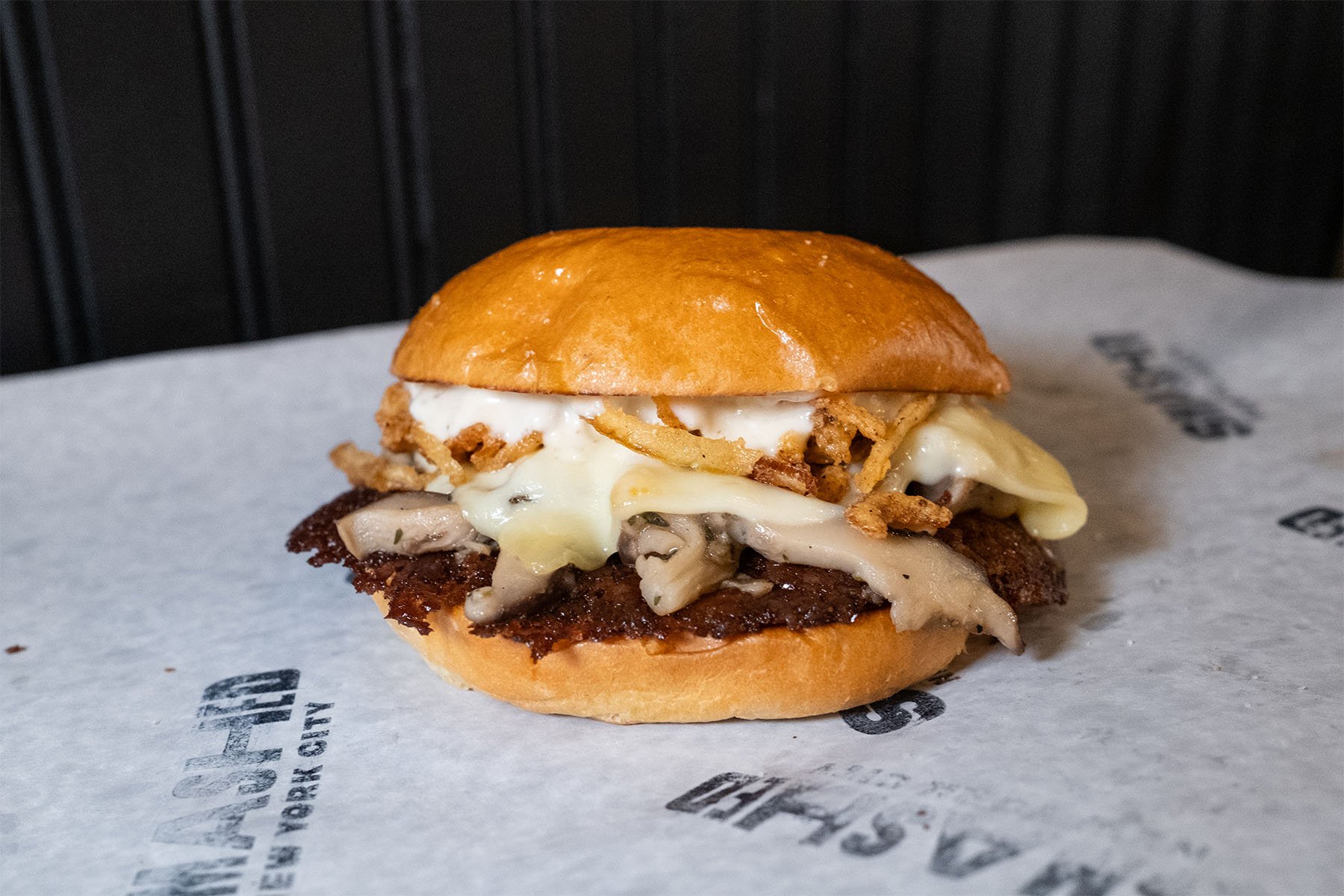 Close-up of a burger with a shiny brioche bun, fried onions, melted cheese, sautéed mushrooms, a grilled beef patty, all sitting on branded paper.