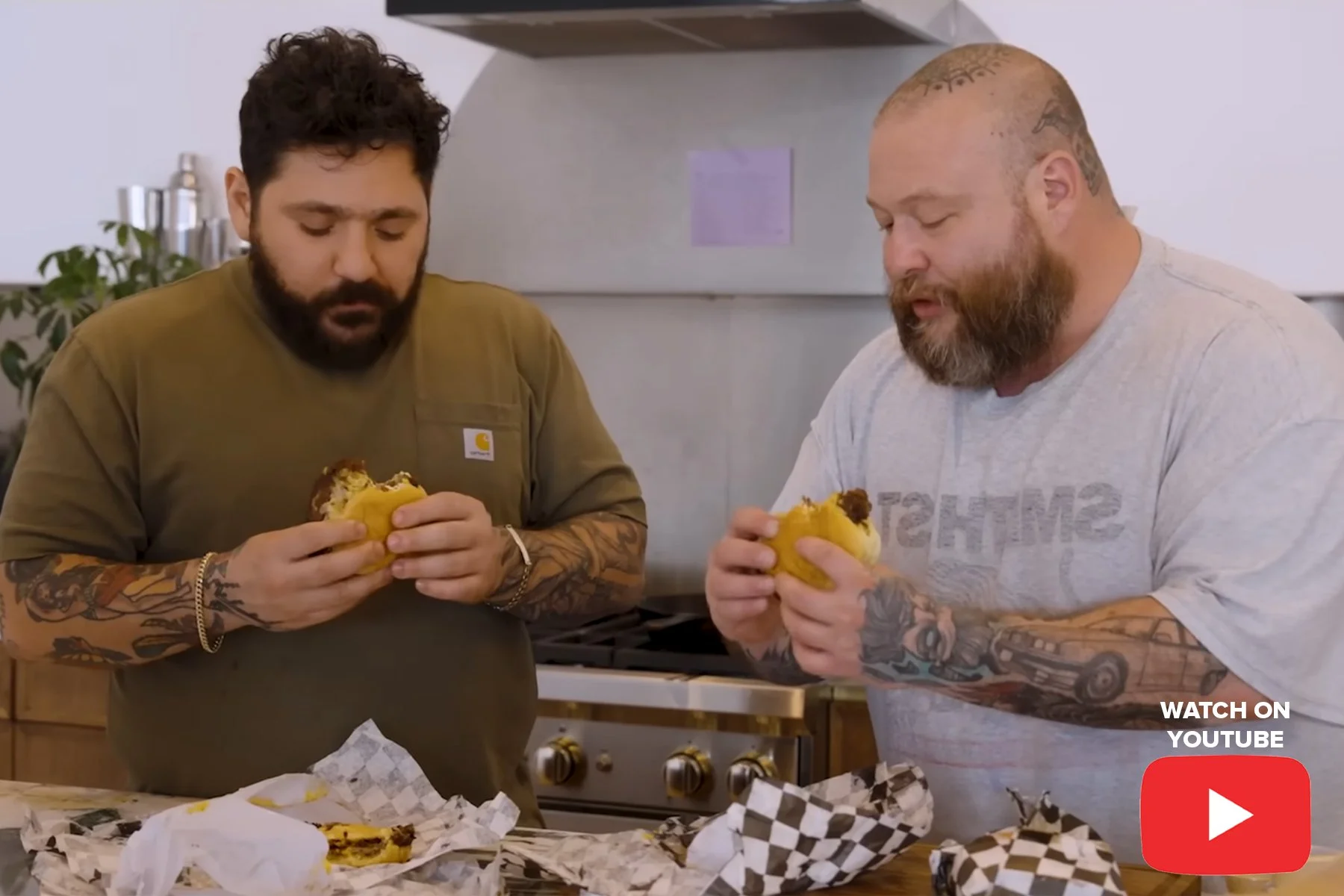 Two tattooed men with beards in a kitchen, holding sandwiches and examining their contents. There are wrappers and food on the counter in front of them.