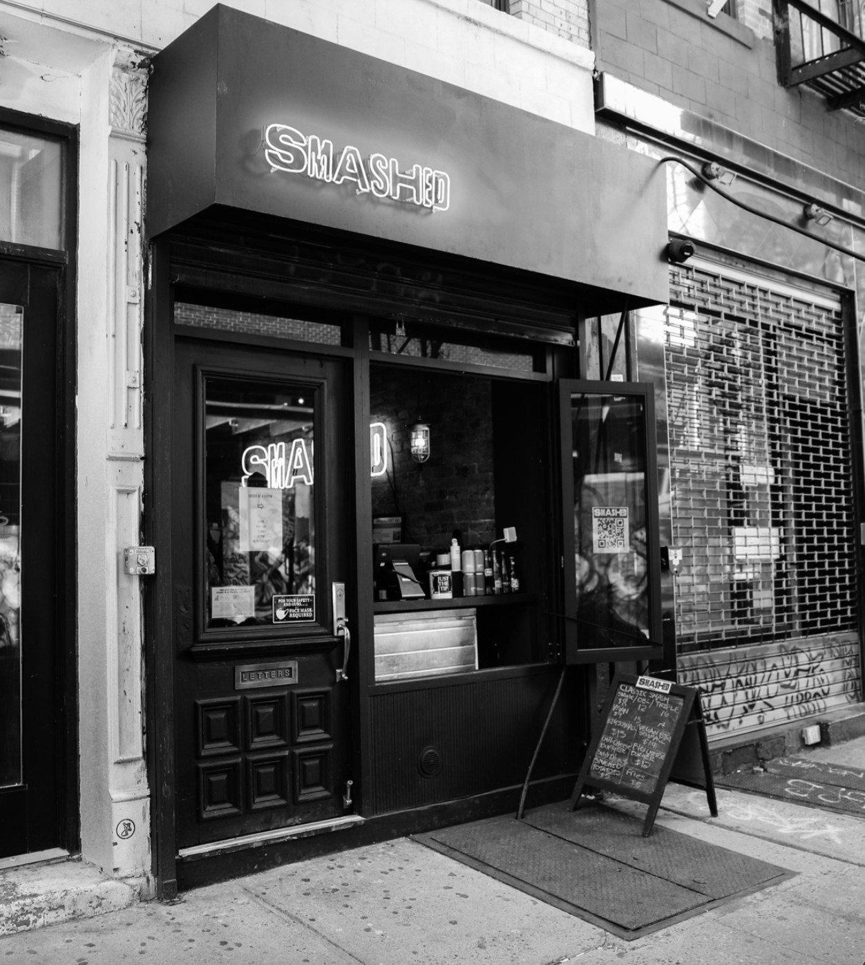 Black and white photo of a small storefront with a neon sign reading 'SMASH' above the entrance. The door has a small window and a sign with a menu board outside. The window displays a neon 'SMASH' sign inside, and the building has brick walls and a trash can nearby.