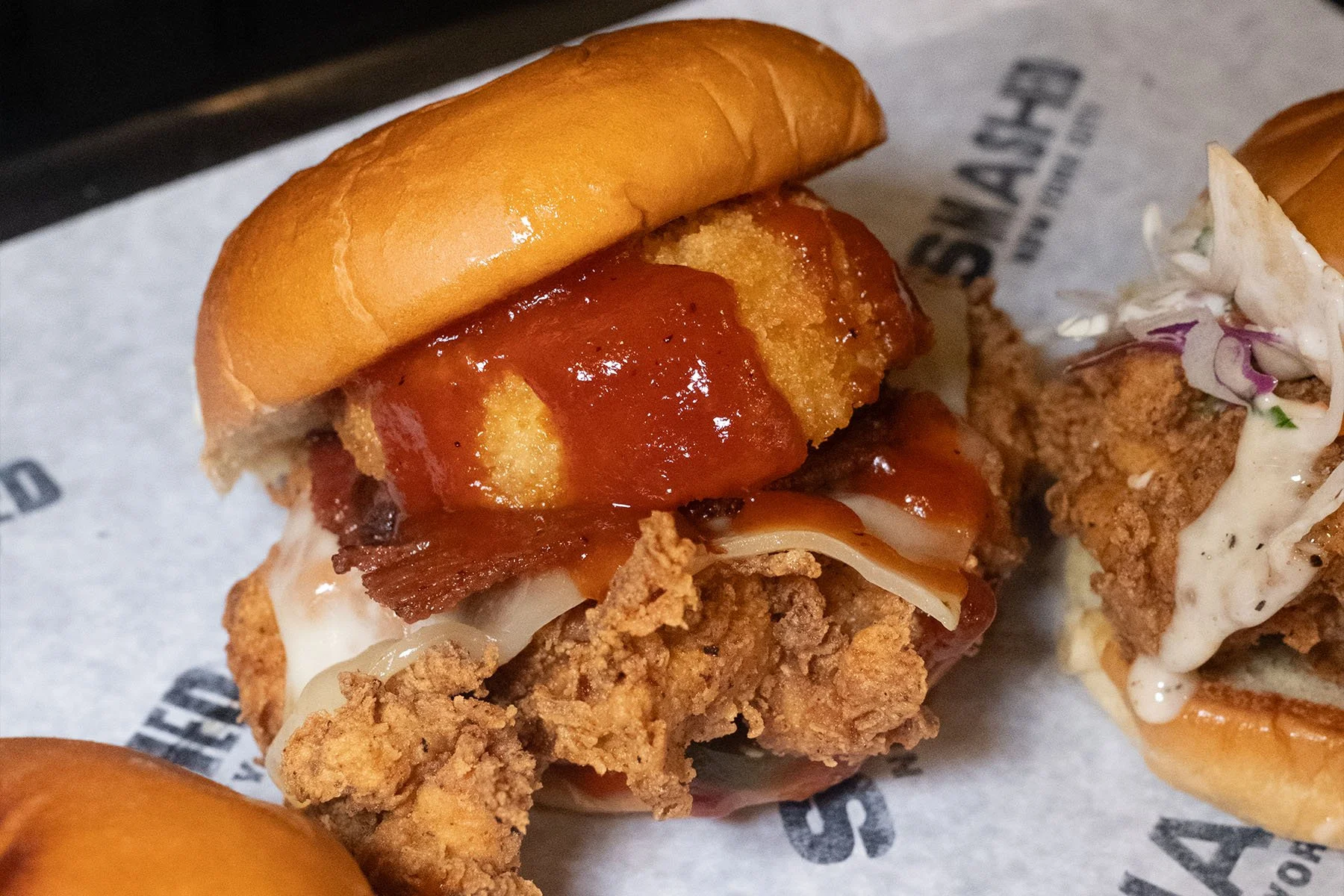 Close-up of a fried chicken sandwich with crispy chicken, bacon, lettuce, sauce, and a bun, on a sheet of parchment paper.