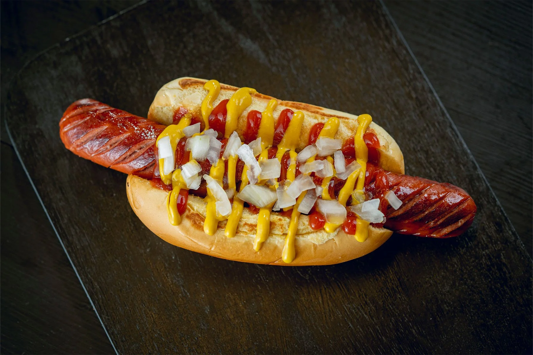 Hot dog in a bun with grilled sausage, mustard, ketchup, and chopped onions on a black plate.