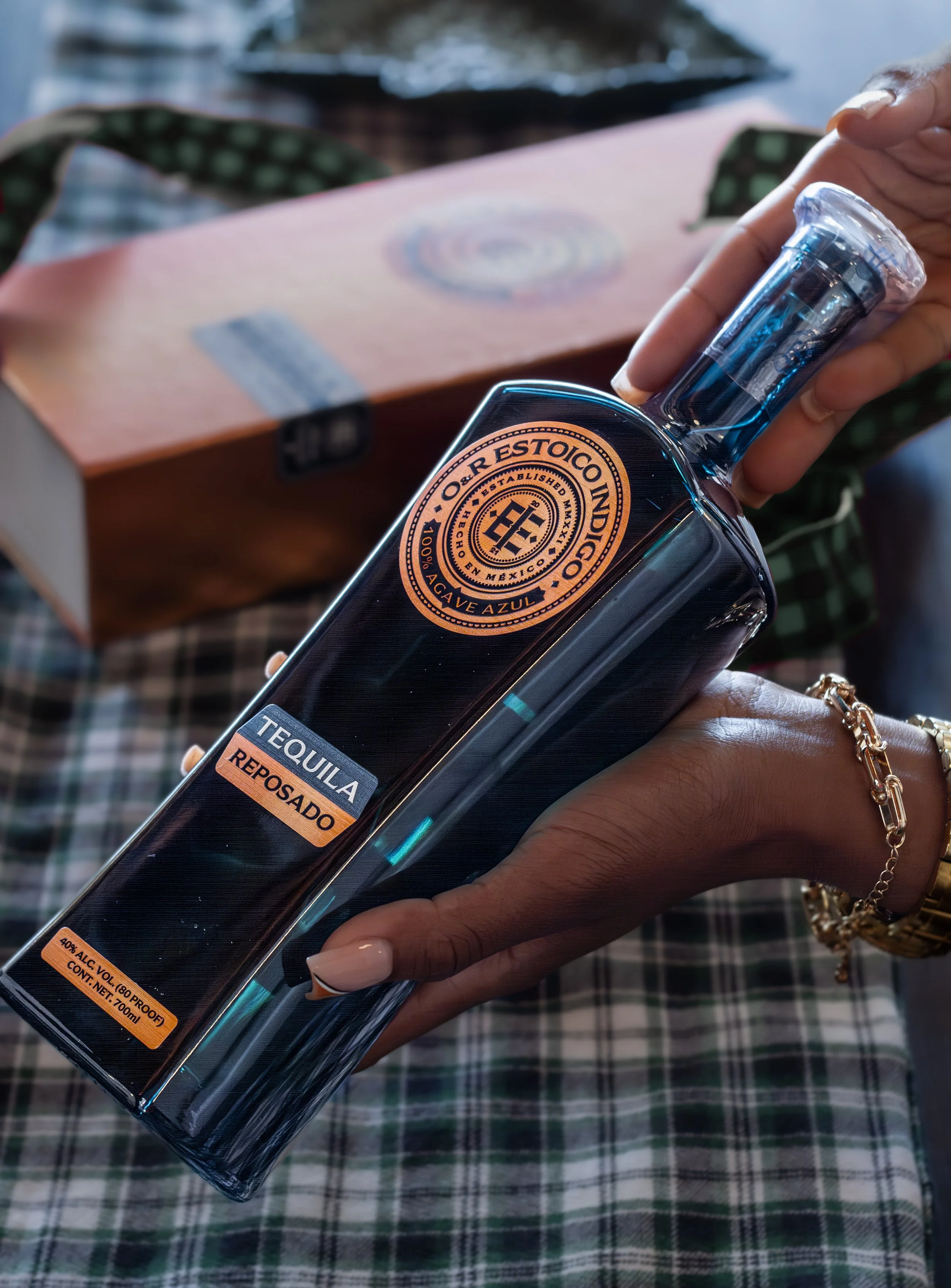 A hand holding a bottle of tequila reposado with a black and orange label. The label includes the text 'O. R. ESTOICO NDI.C.O', 'AGAVE AZUL', 'TEQUILA REPOSADO', and alcohol content information. The person is wearing a gold bracelet and has light-colored nails. In the background, there is a wooden box and a checkered cloth.