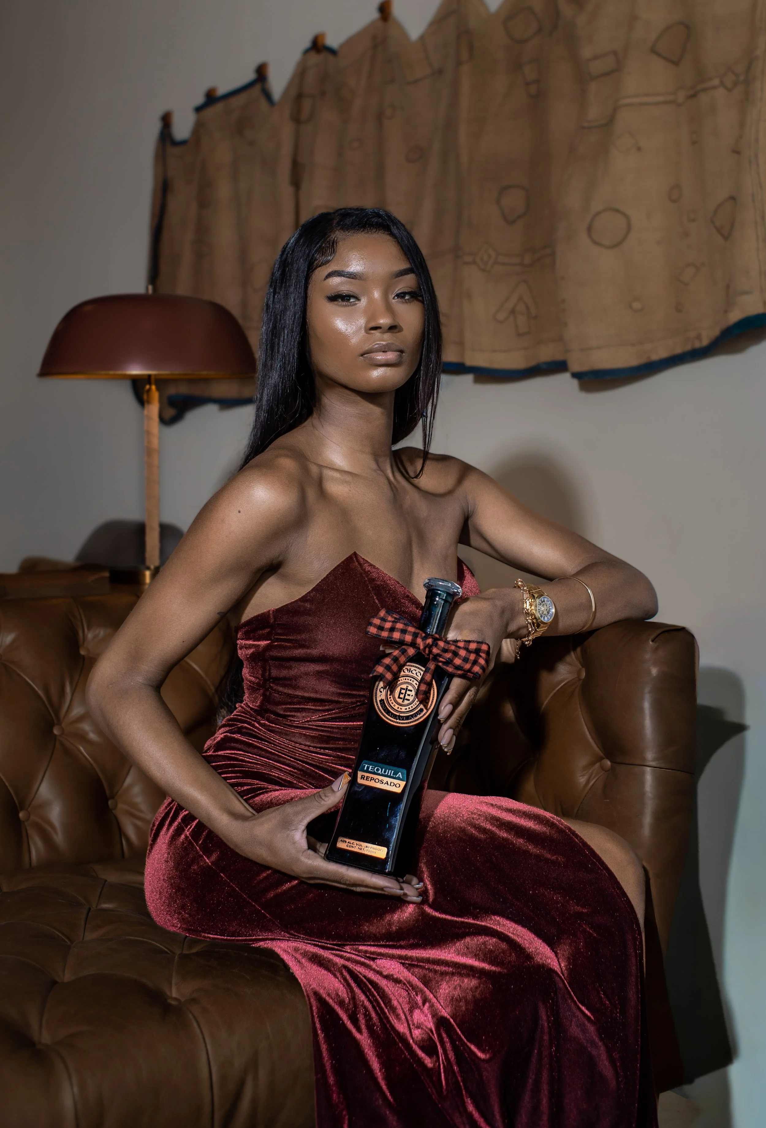 A woman in a strapless velvet dress sitting on a brown leather couch, holding a bottle of Tequila Reposado with a red and black checkered bow. She has long black hair, gold jewelry, and a watch. The background features a curtain and a lamp.