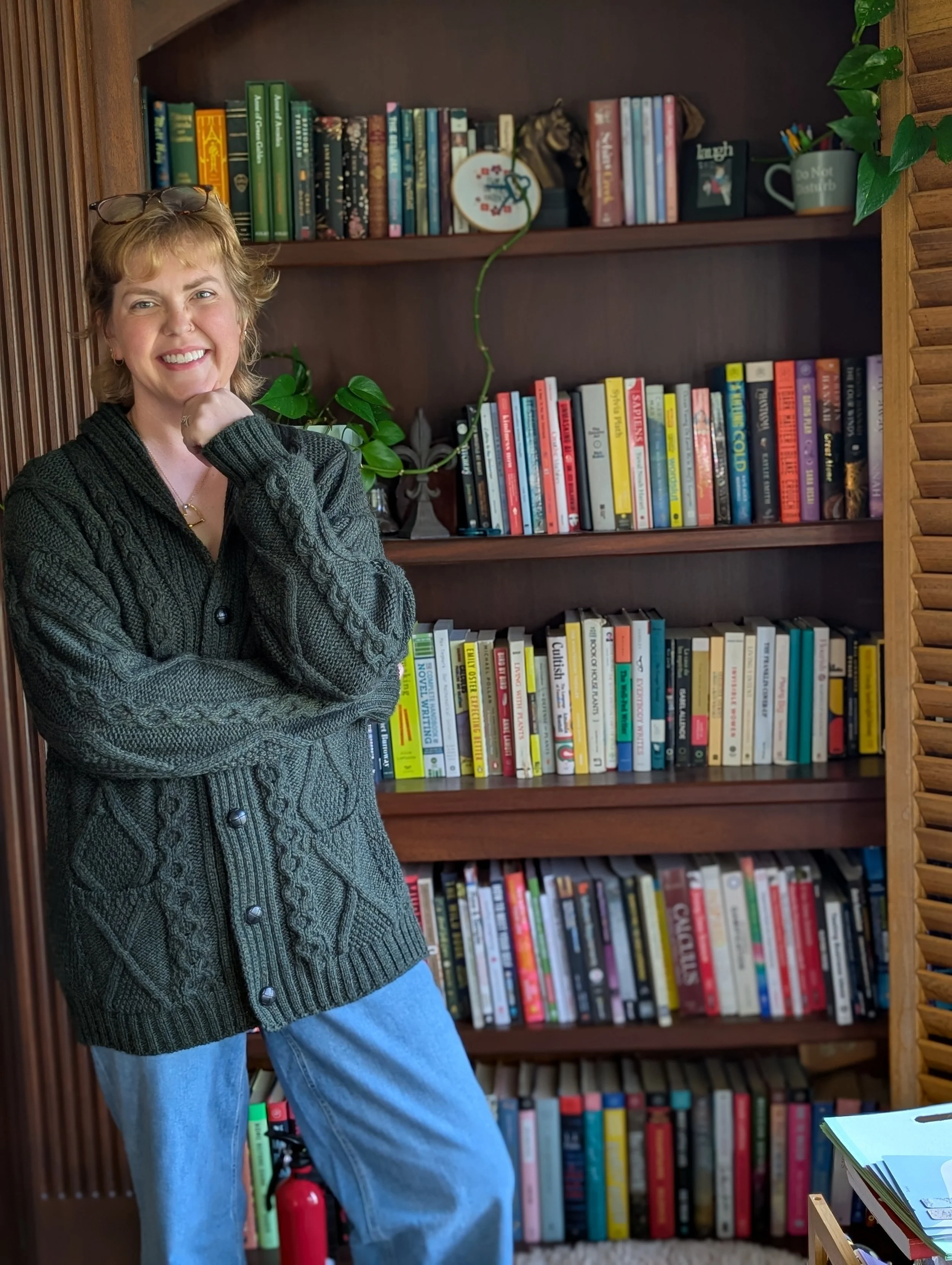 Smile of a woman wearing a dark green cardigan and blue jeans, standing in front of a wooden bookshelf filled with colorful books and some potted plants.