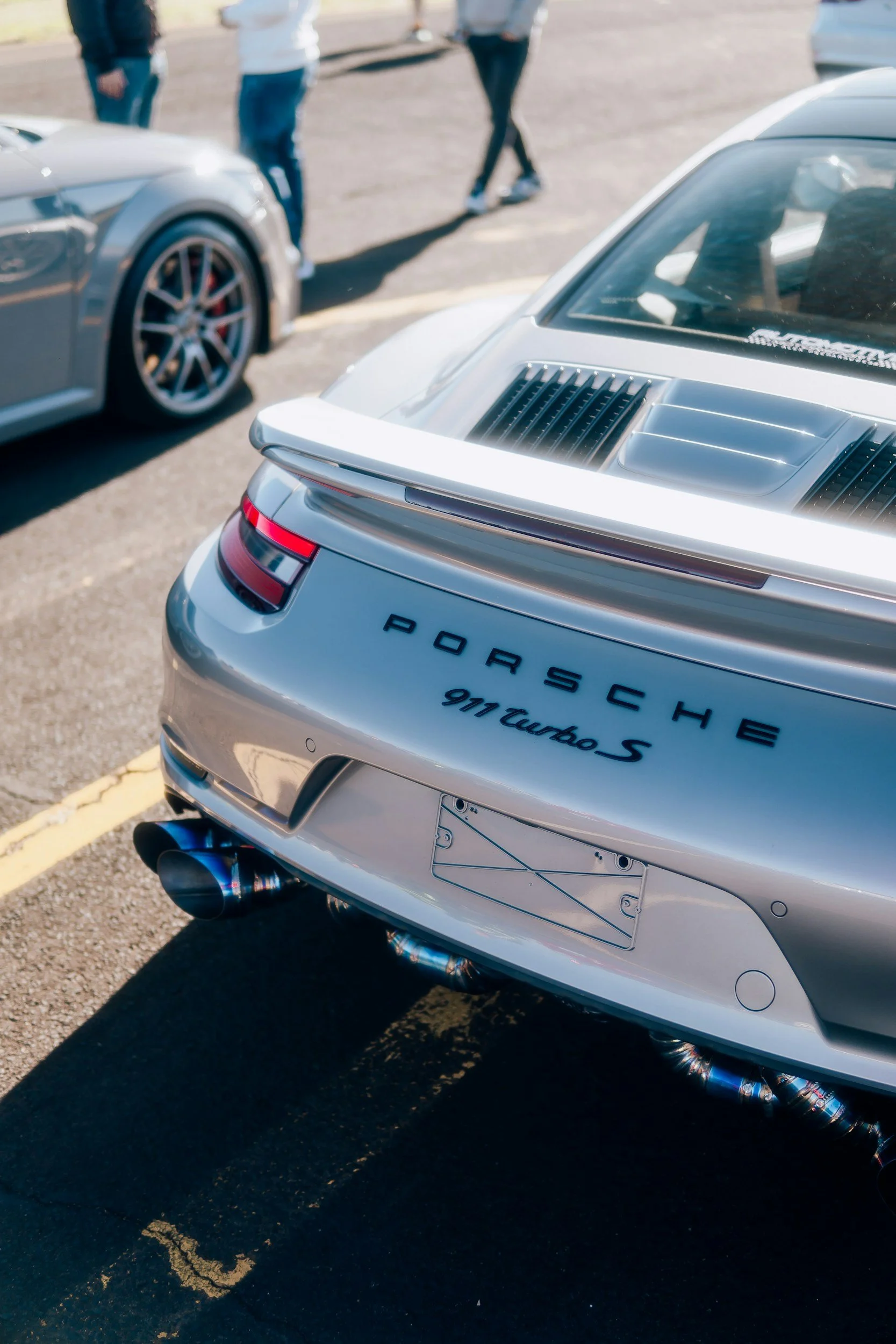 Rear view of a silver Porsche 911 Turbo S parked in a lot, with another car and people walking nearby.