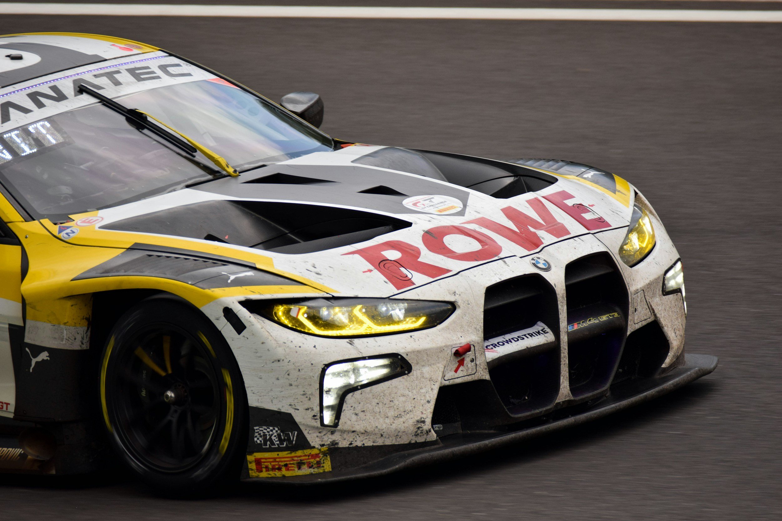 A racing car on the track with dirt and tire marks on its front bumper and body, featuring a white, yellow, and black color scheme with prominent sponsor logos.