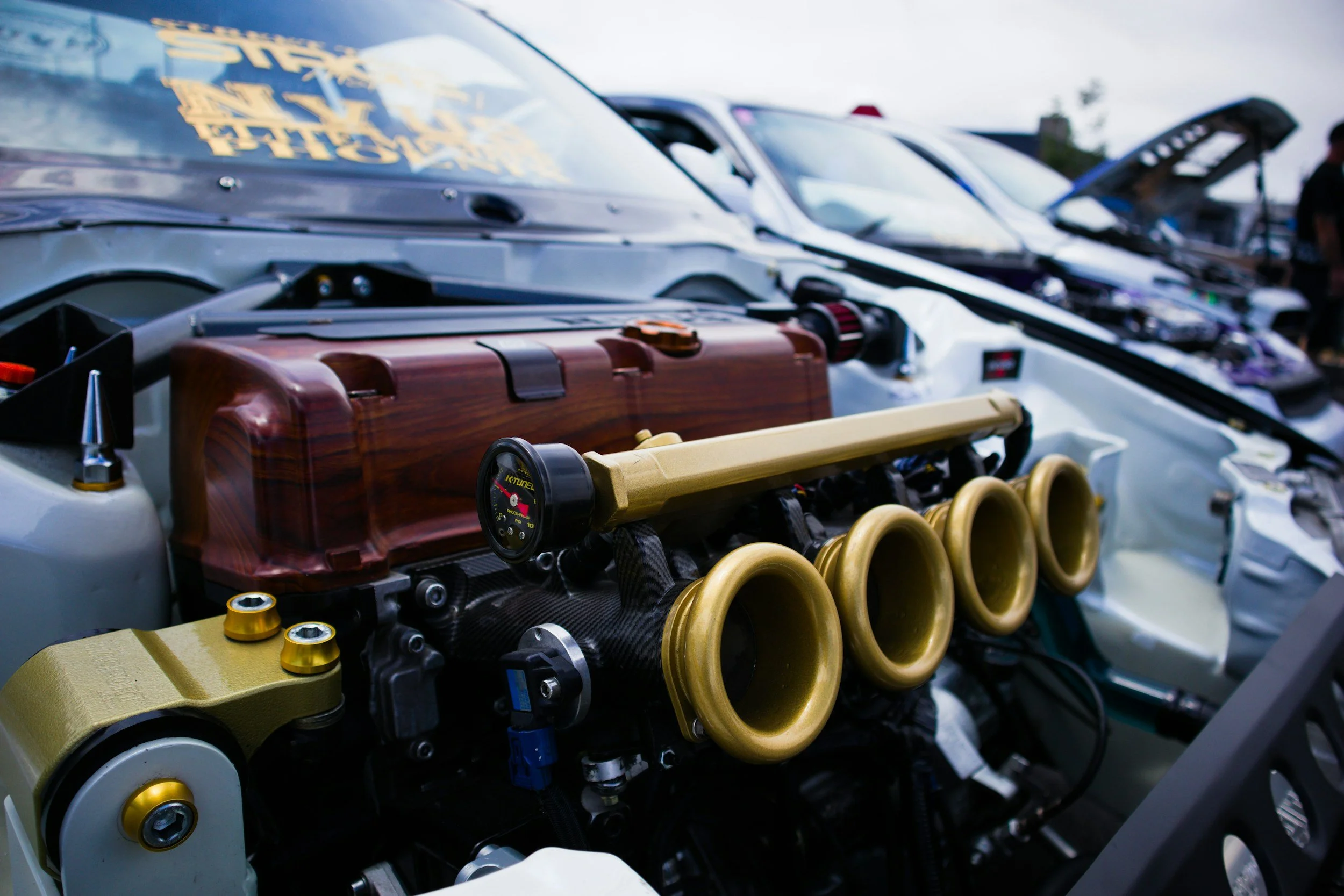 Close-up of a car's engine bay, featuring a polished wooden oil cooler cover, yellow intake trumpets, and various engine components, with other cars visible in the background at a car show.