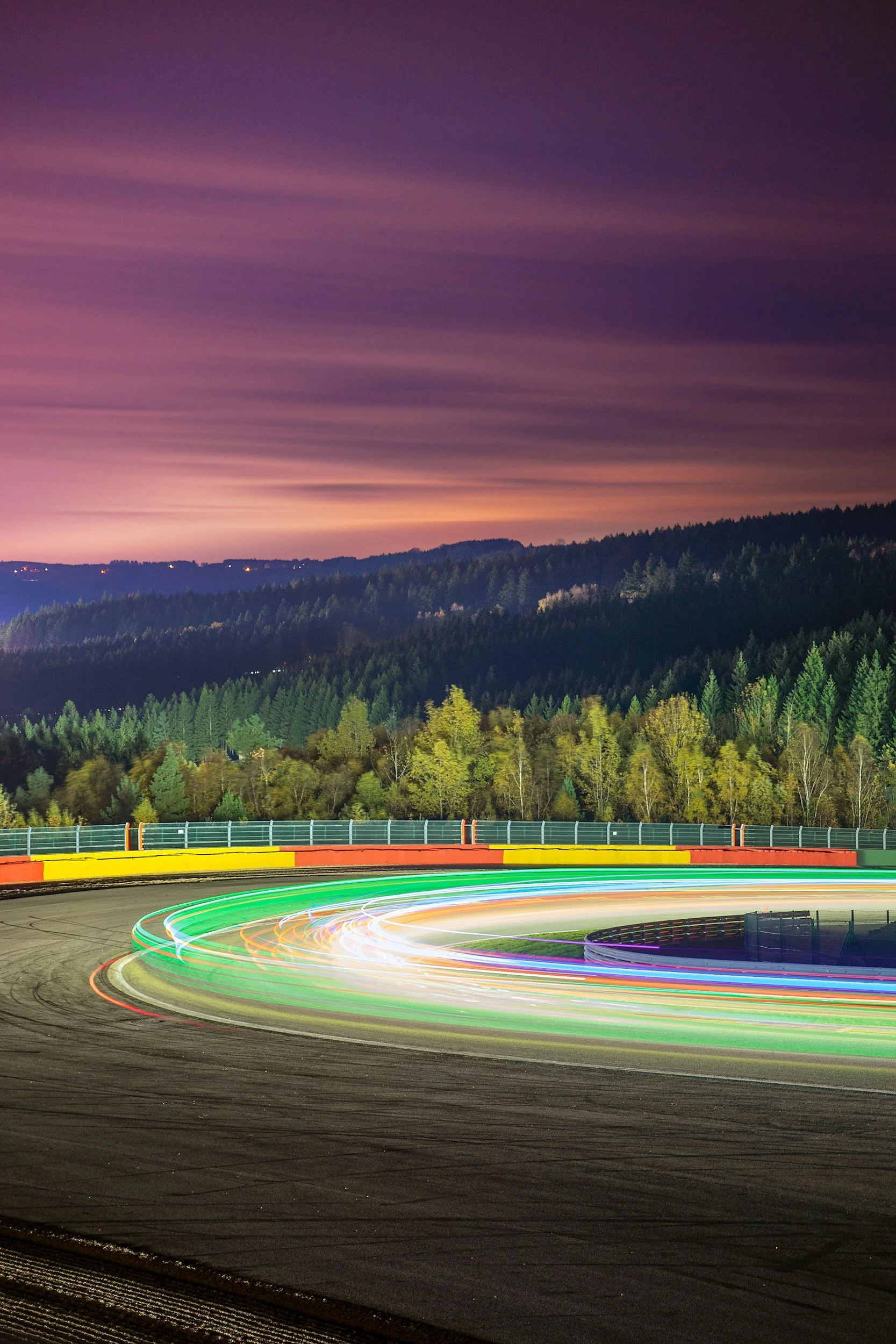 Light trails from vehicles racing on a winding dirt race track at night, with a forested hillside and colorful sky in the background.