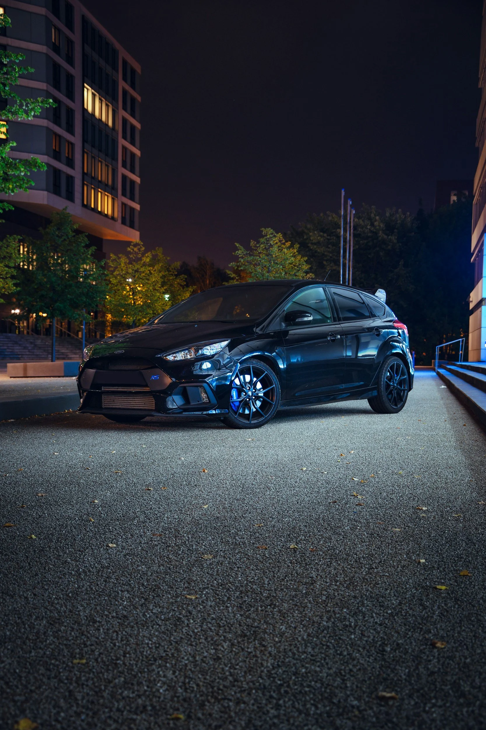 A black sports hatchback car parked on a city street at night with lit buildings and trees in the background.