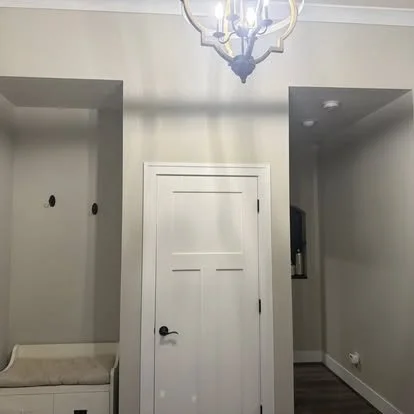 Interior view of a room featuring a central white door, a chandelier hanging from the ceiling, and a small bench or cabinet to the left.