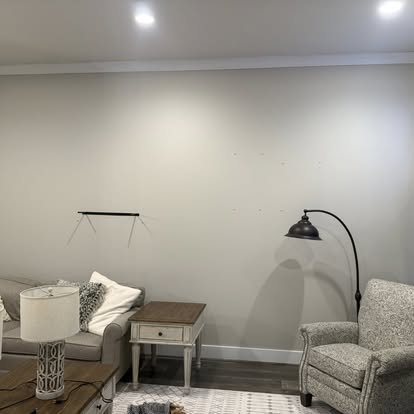 A living room with a beige sofa, a white side table, a gray armchair, a black floor lamp, and a small wall shelf on a gray wall.