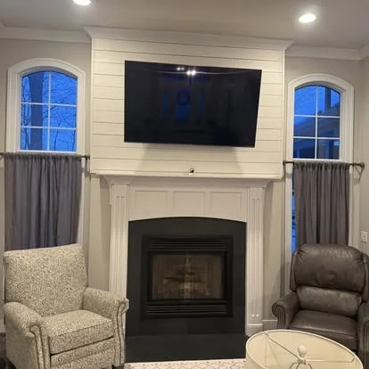 Living room with fireplace, two windows with curtains, and a wall-mounted TV above the fireplace.