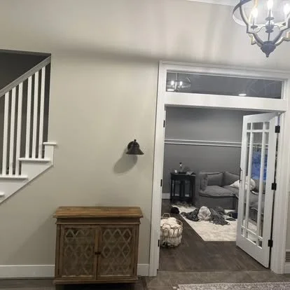 Living room seen through open French doors with a gray sofa, coffee table, and dog lying on the floor. A wooden cabinet is positioned against a wall, and a black wall-mounted light fixture is above it. Part of a staircase with white spindles is visib