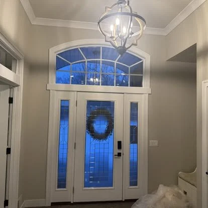 Front door with a Christmas wreath, side windows, and blue-tinted glass panels under a large arched window, inside a home with a chandelier hanging from the ceiling.