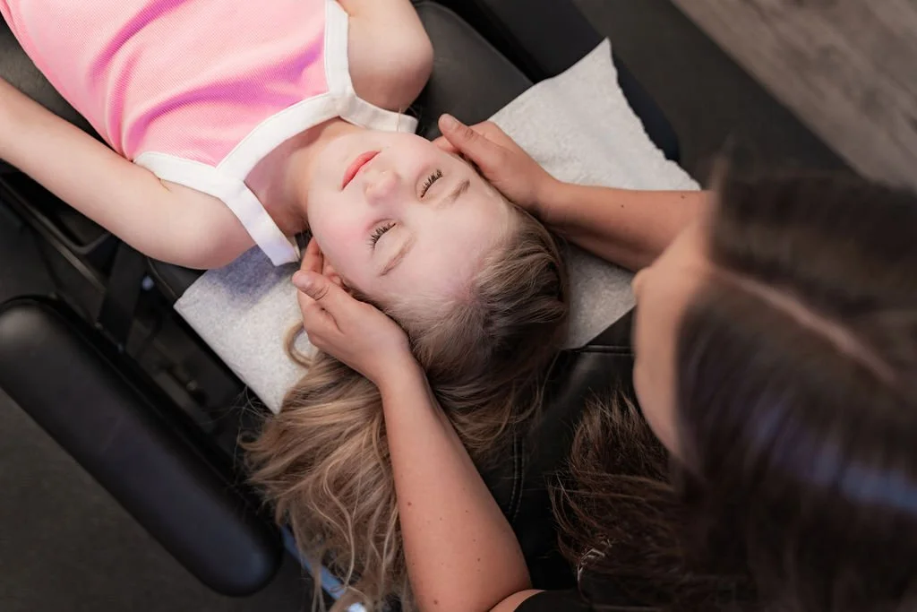A young girl with light brown hair lying face up on a black treatment table, receiving a chiropractic adjustment while a practitioner gently holds her head.