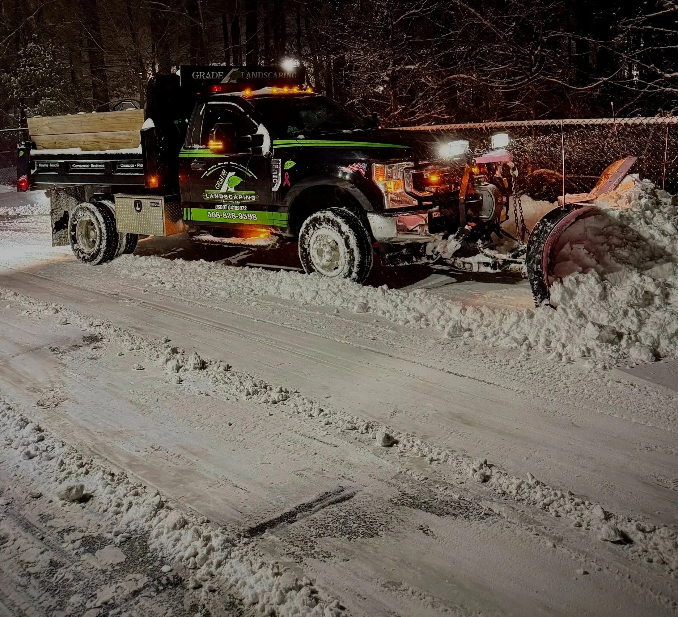 A snowplow truck clearing snow from a road during nighttime, with snow-covered trees in the background.