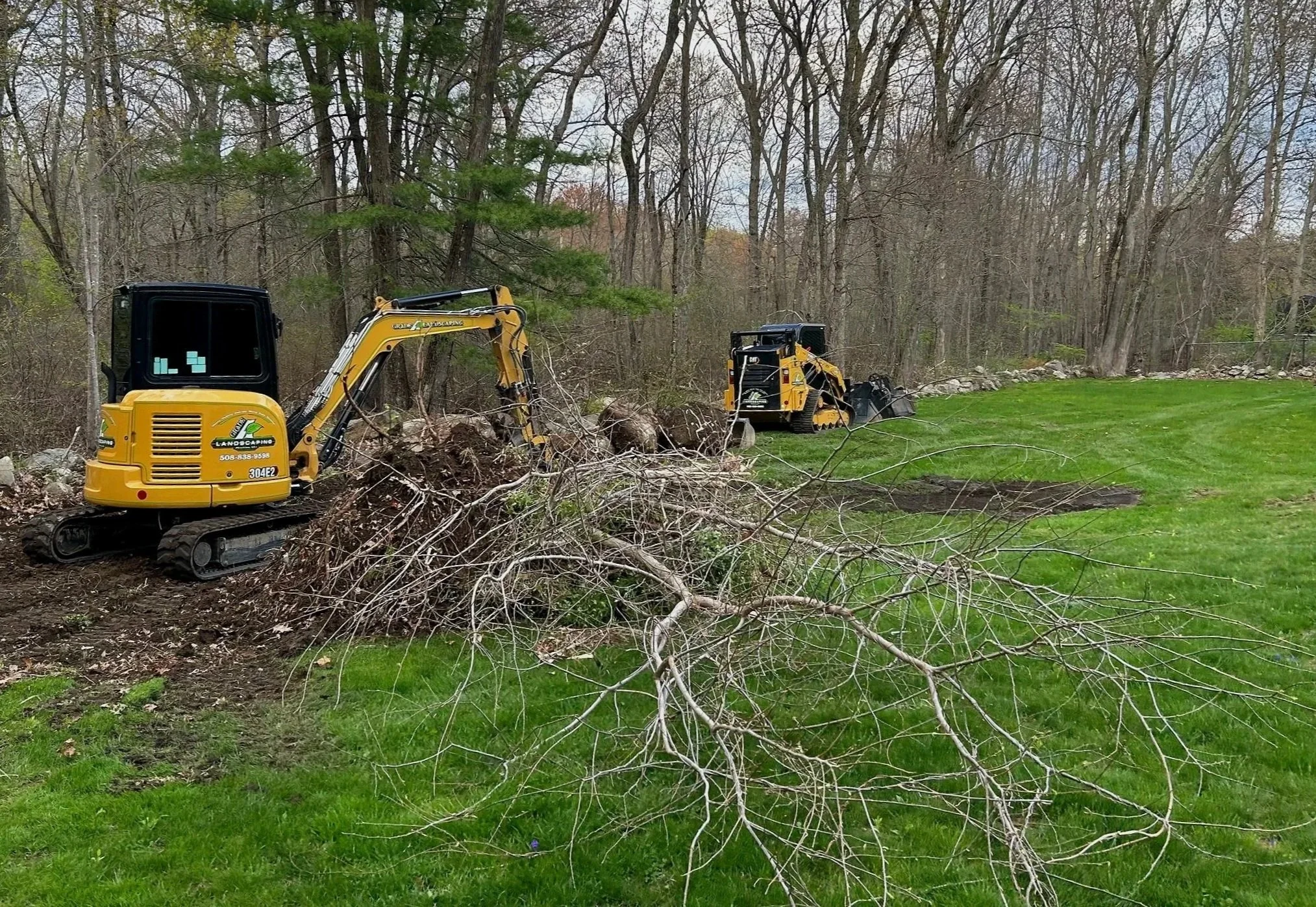 Two yellow landscaping mini excavators working on removing tree branches in a backyard with green grass and trees in the background.
