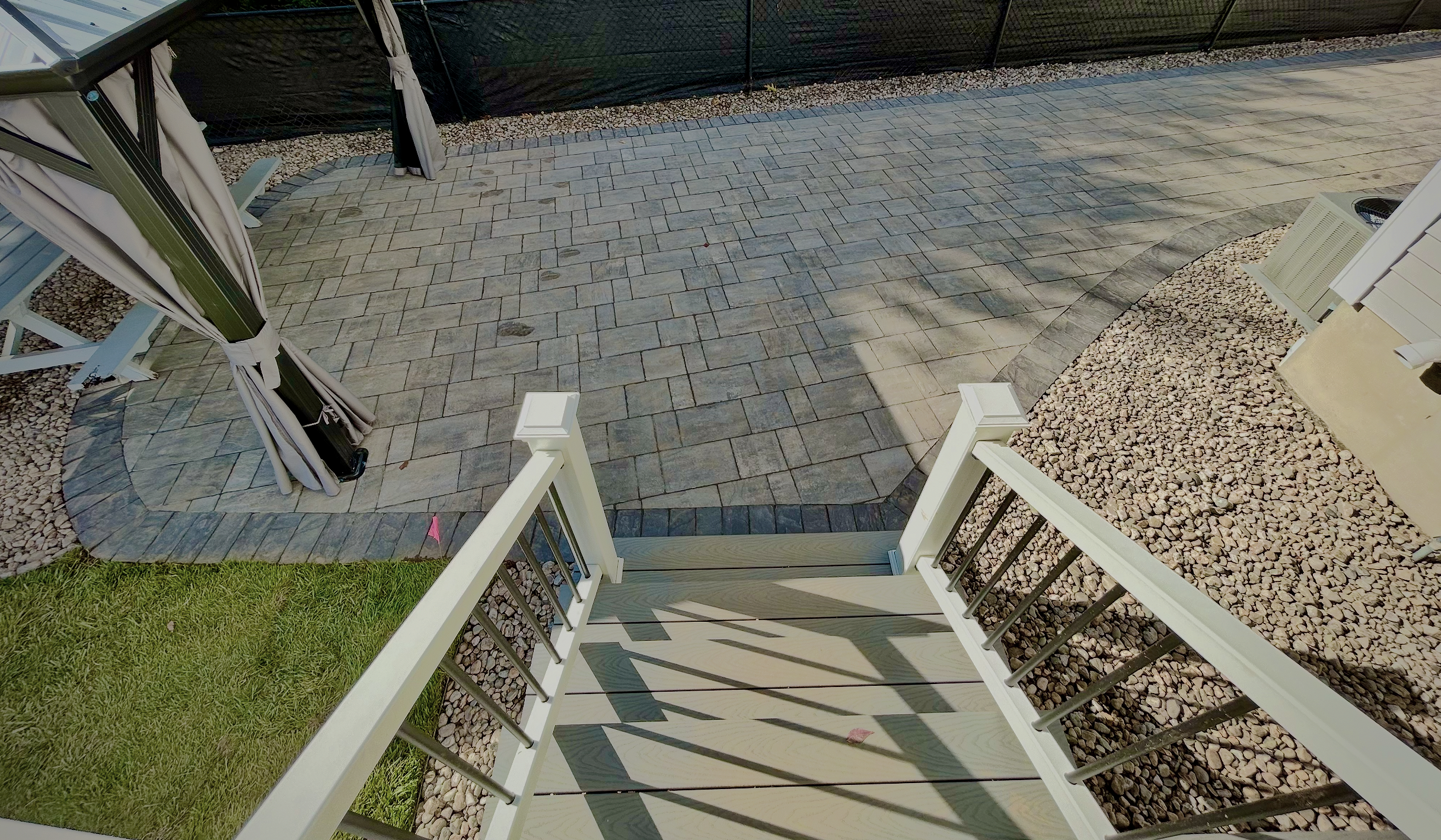 View from a staircase looking down on a backyard patio area with paving stones, gravel, and a small patch of grass, with an outdoor umbrella folded beside the house.