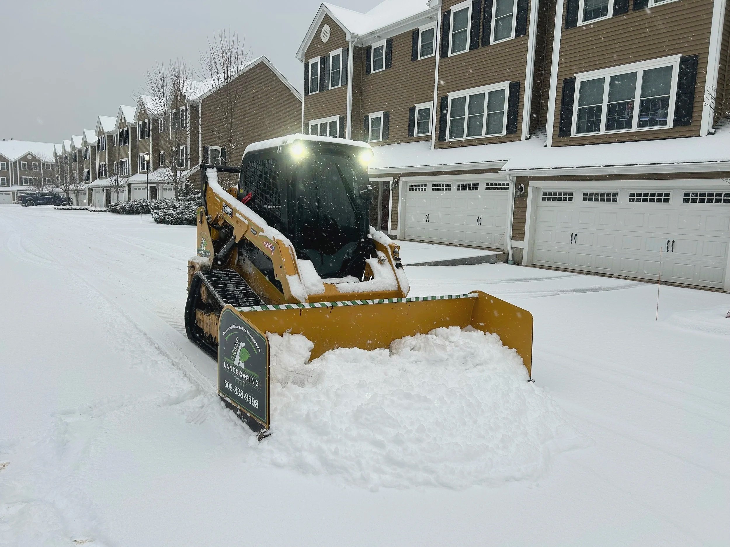 A skid-steer loader clearing snow from a residential street with townhouses in the background.