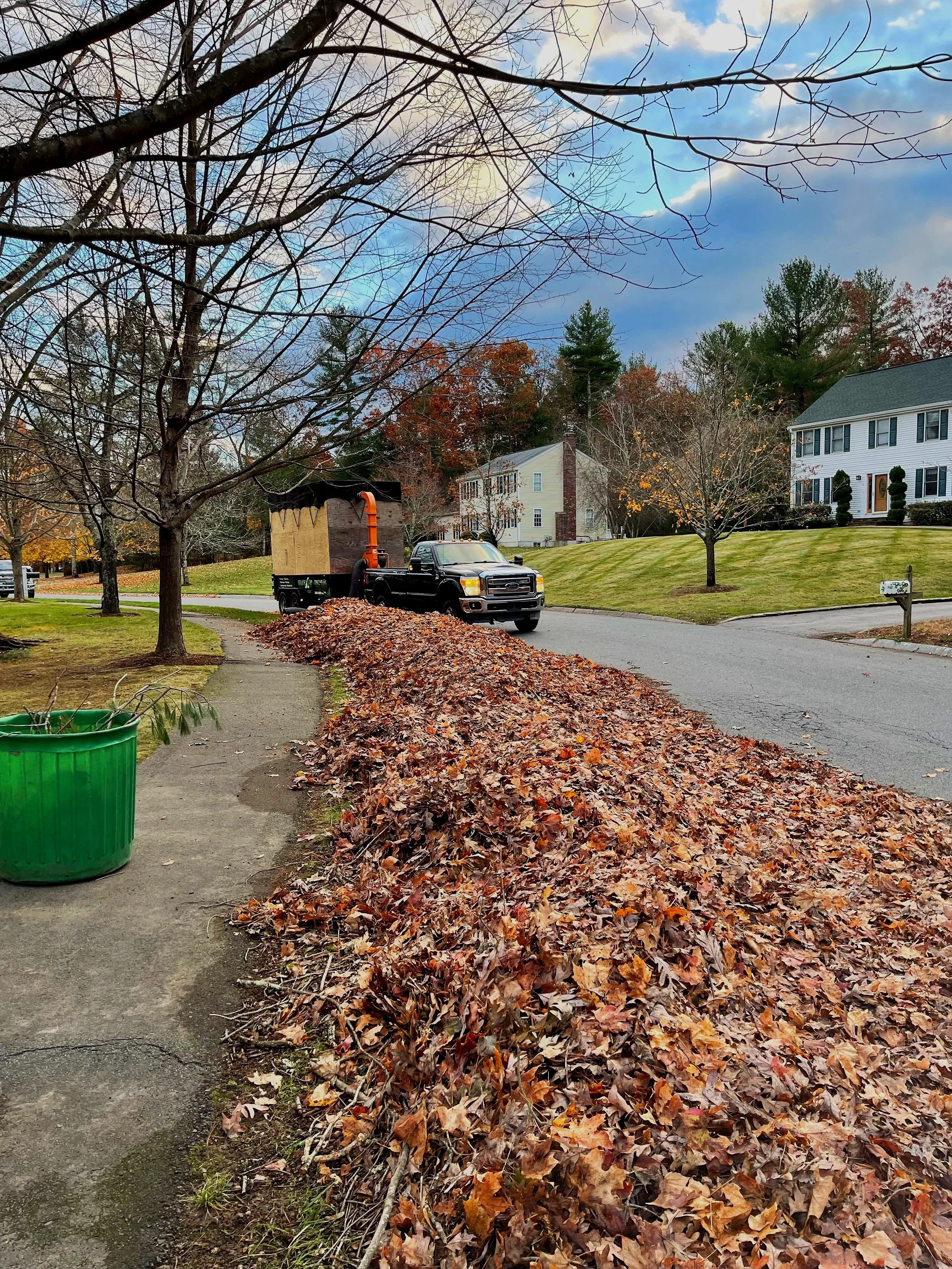 A truck with a leaf vacuum attachment collecting fallen leaves along the curb of a suburban street during autumn. The street is lined with trees, some with remaining leaves and others bare, and residential houses in the background.