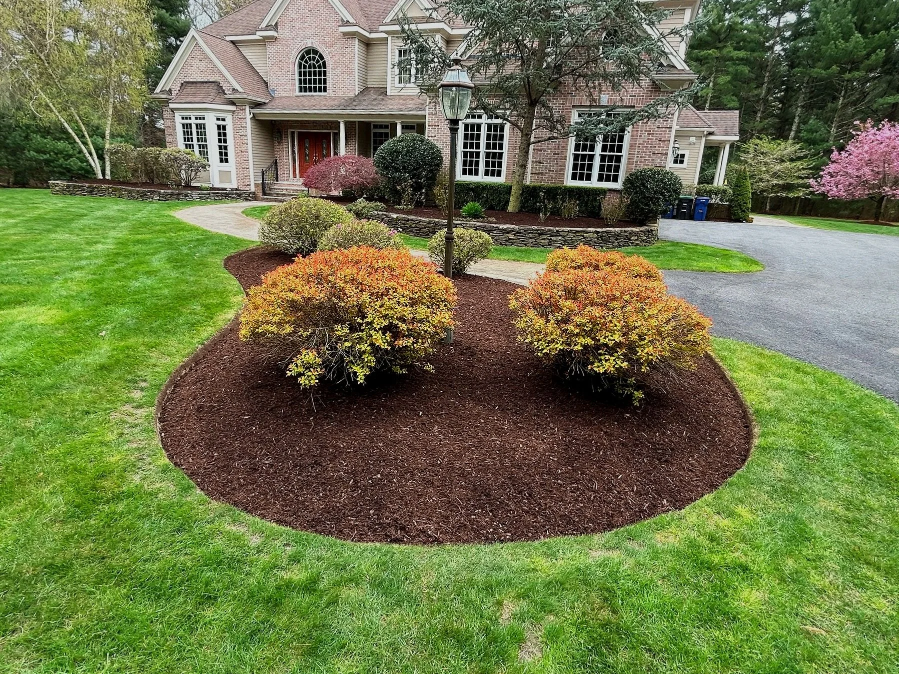 A landscaped front yard with a curved garden bed filled with bushes, mulch, and a lamppost in front of a large brick house with a rounded driveway.