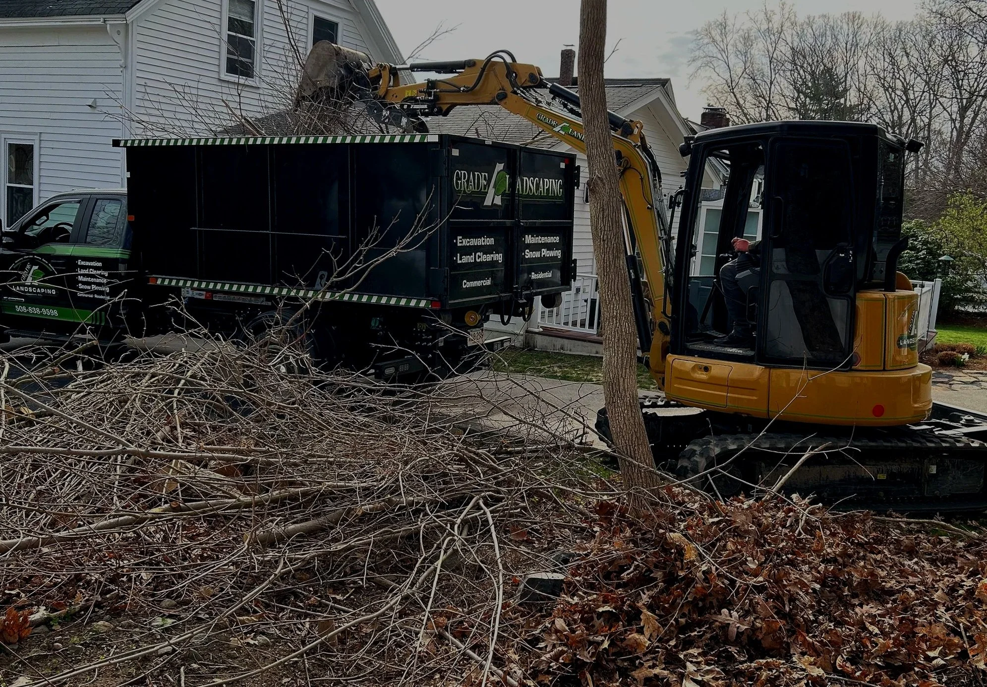 A small yellow excavator removing tree branches and leaves from a yard, with a black landscaping truck nearby, in front of a white house.