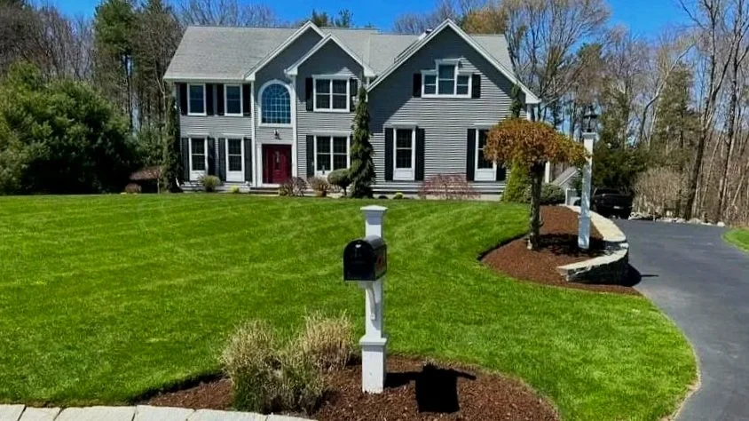 A large two-story house with gray siding and multiple windows, surrounded by a well-maintained green lawn, small trees, and a curved driveway.