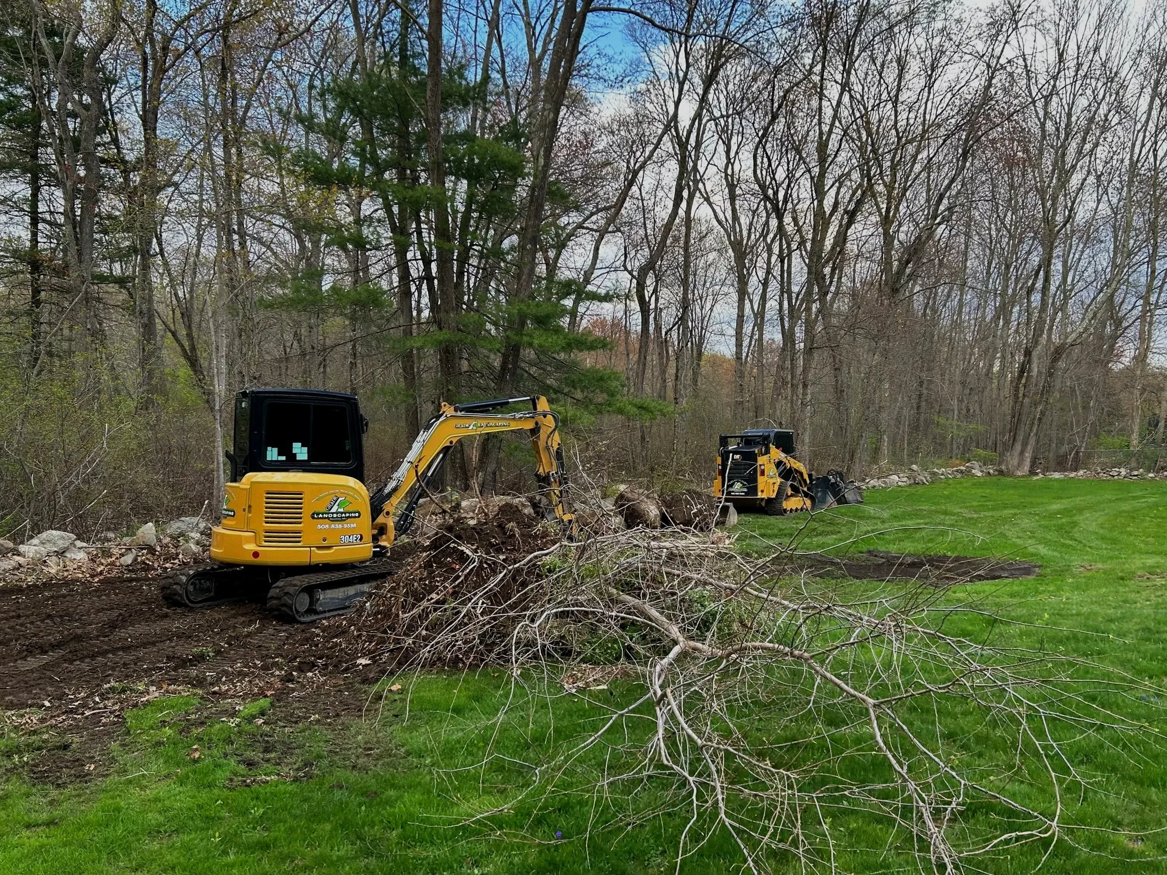 Two yellow mini excavators working on clearing fallen branches from a wooded area next to a well-maintained grassy lawn.