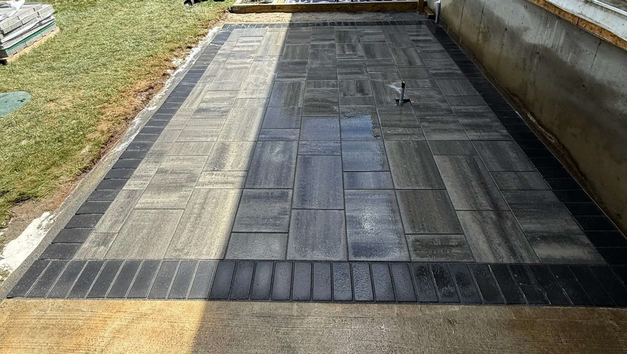 Newly installed grey and black paver walkway outside, with grass on the left and a concrete wall on the right.