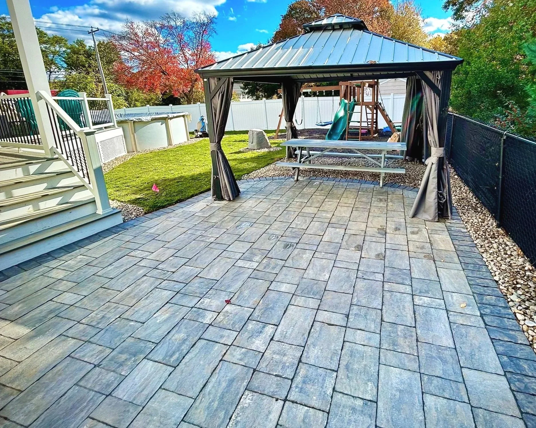 A backyard patio with interlocking stone tiles, a black metal gazebo with curtains, a picnic table, and a children's playset with a slide, swings, and a climbing wall. There is a grassy area, a small above-ground pool, and trees with fall foliage in 