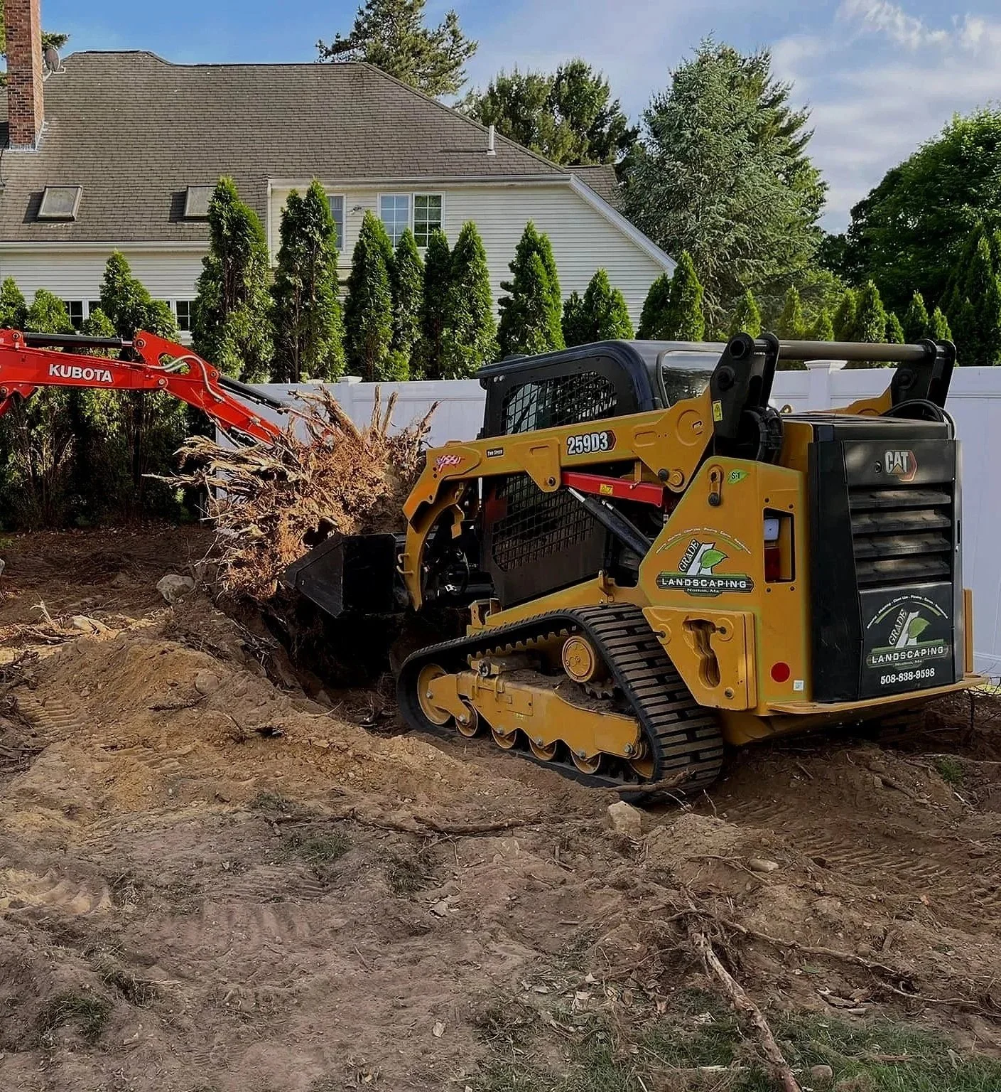 A small yellow bulldozer moving dirt in a backyard with a white fence, trees, and a house in the background.