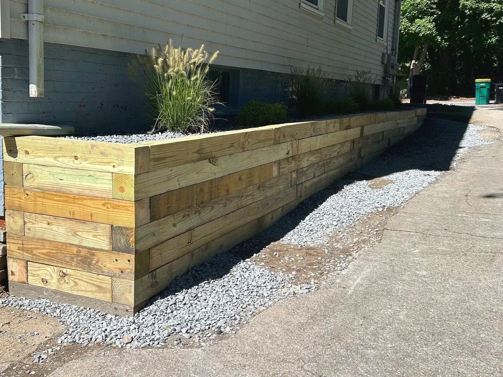 Wooden raised garden bed with plants and gravel lining at the base, next to a house with a beige exterior and a gravel path.