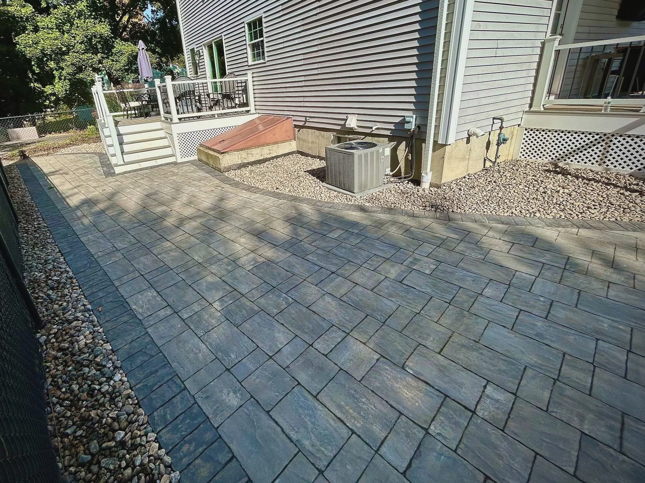 A backyard patio with stone pavers, a fenced deck with outdoor furniture, and a small garden area with gravel close to a house with vinyl siding.
