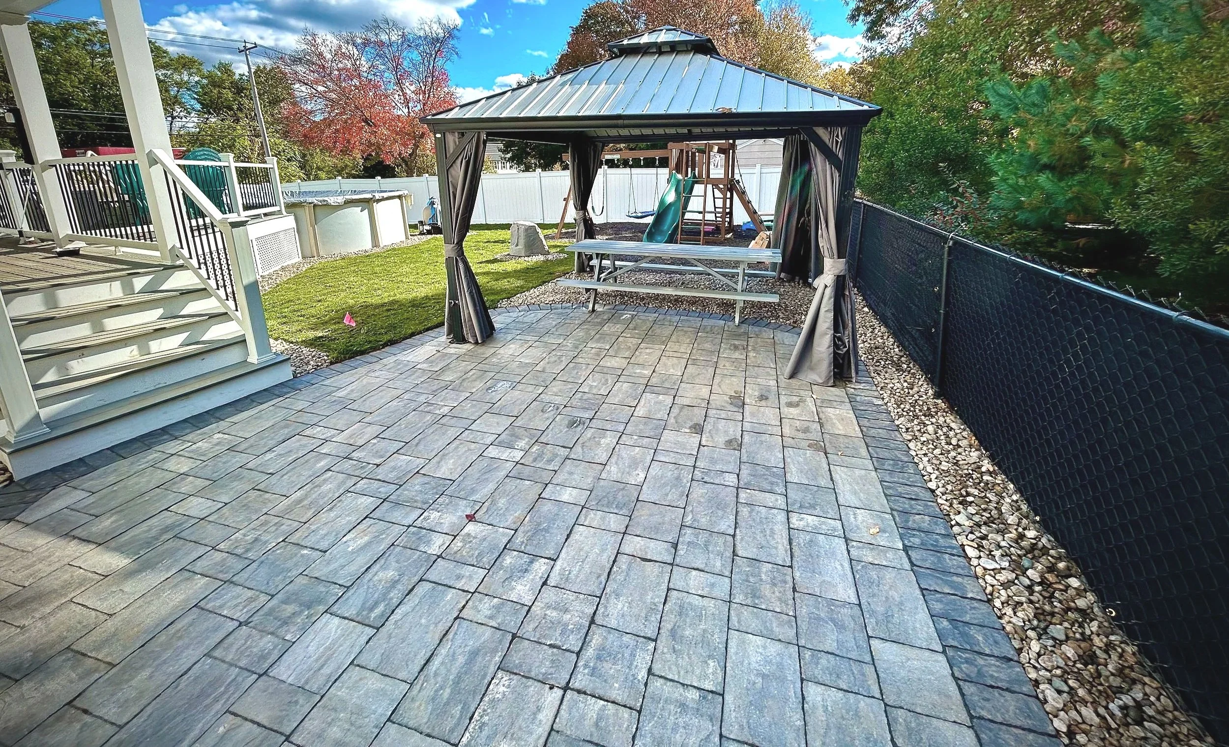 A backyard patio with stone pavers, a gazebo with dark curtains, a wooden picnic table, a children's playset with slide, an above-ground pool, and a white fence with trees in the background.