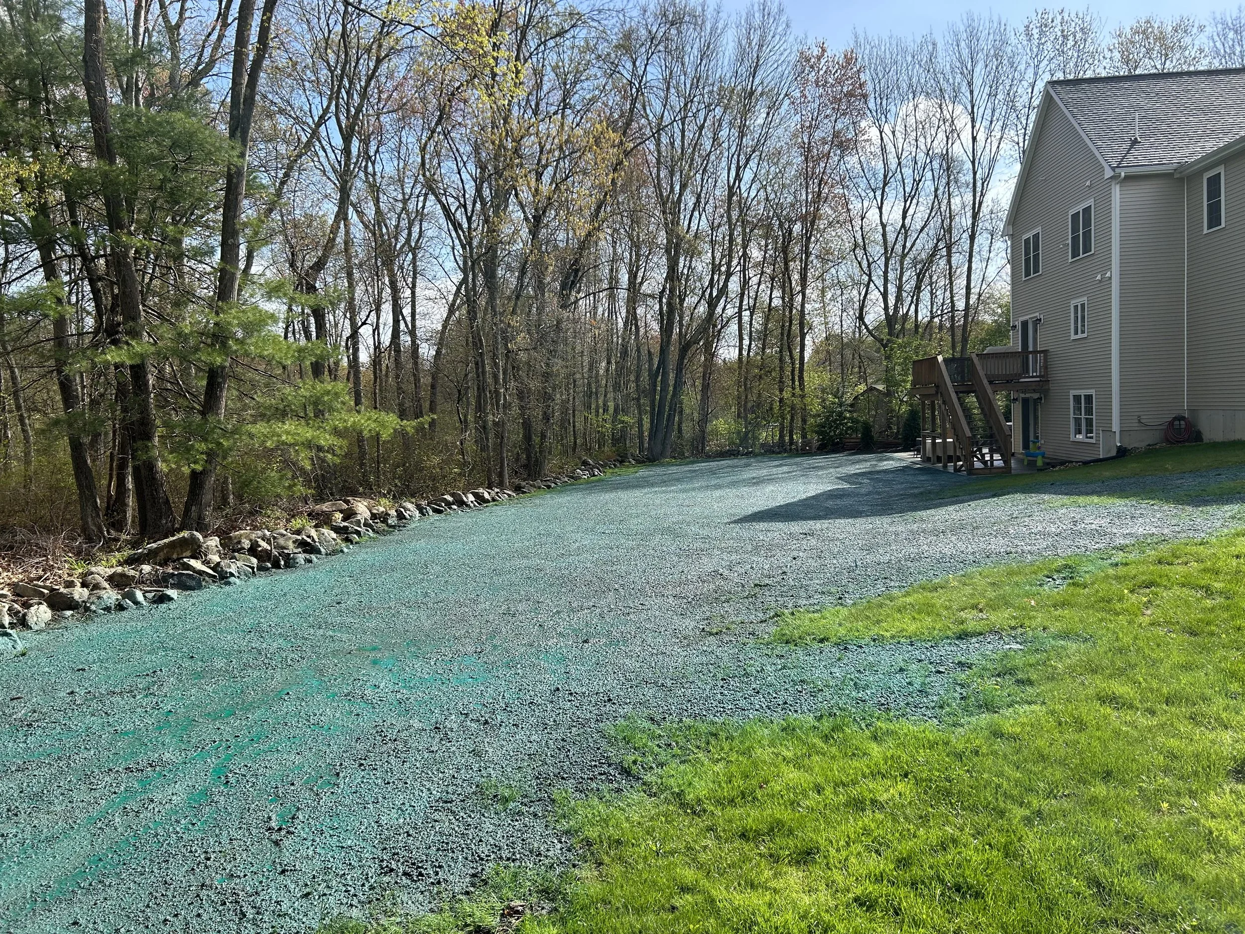 A backyard with a house, gravel driveway, green grass, and trees along the edge of the yard.