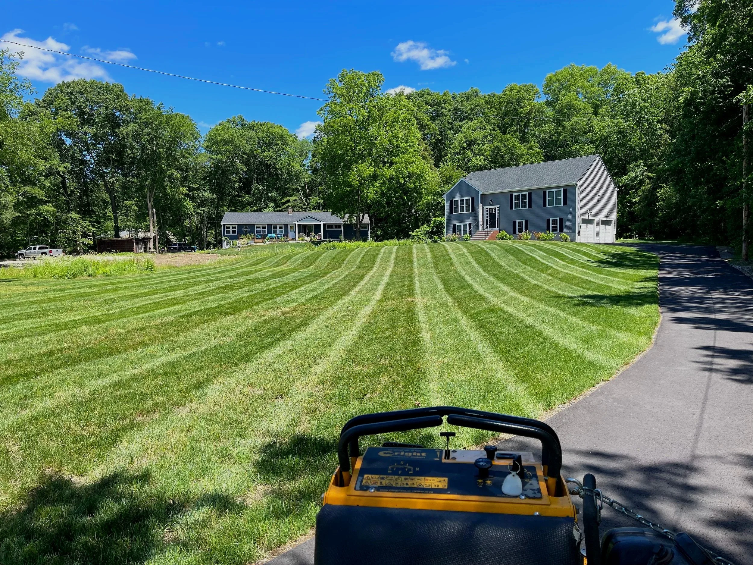 A well-maintained lawn with freshly mowed green grass, striped pattern, and two houses in the background under a clear blue sky with a few clouds. A driveway runs along the right side, and a lawn mower is visible in the foreground.