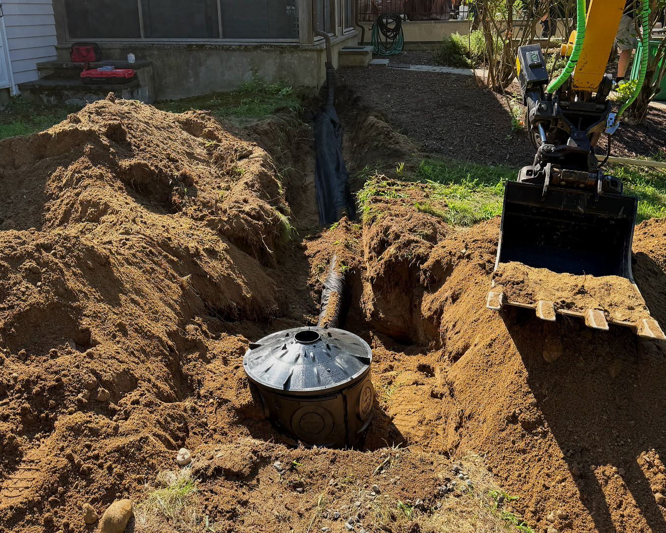 Undergoing underground pipe installation with a trench dug in the yard, an excavator bucket, and a black pipe connected to a septic tank or utility access point.