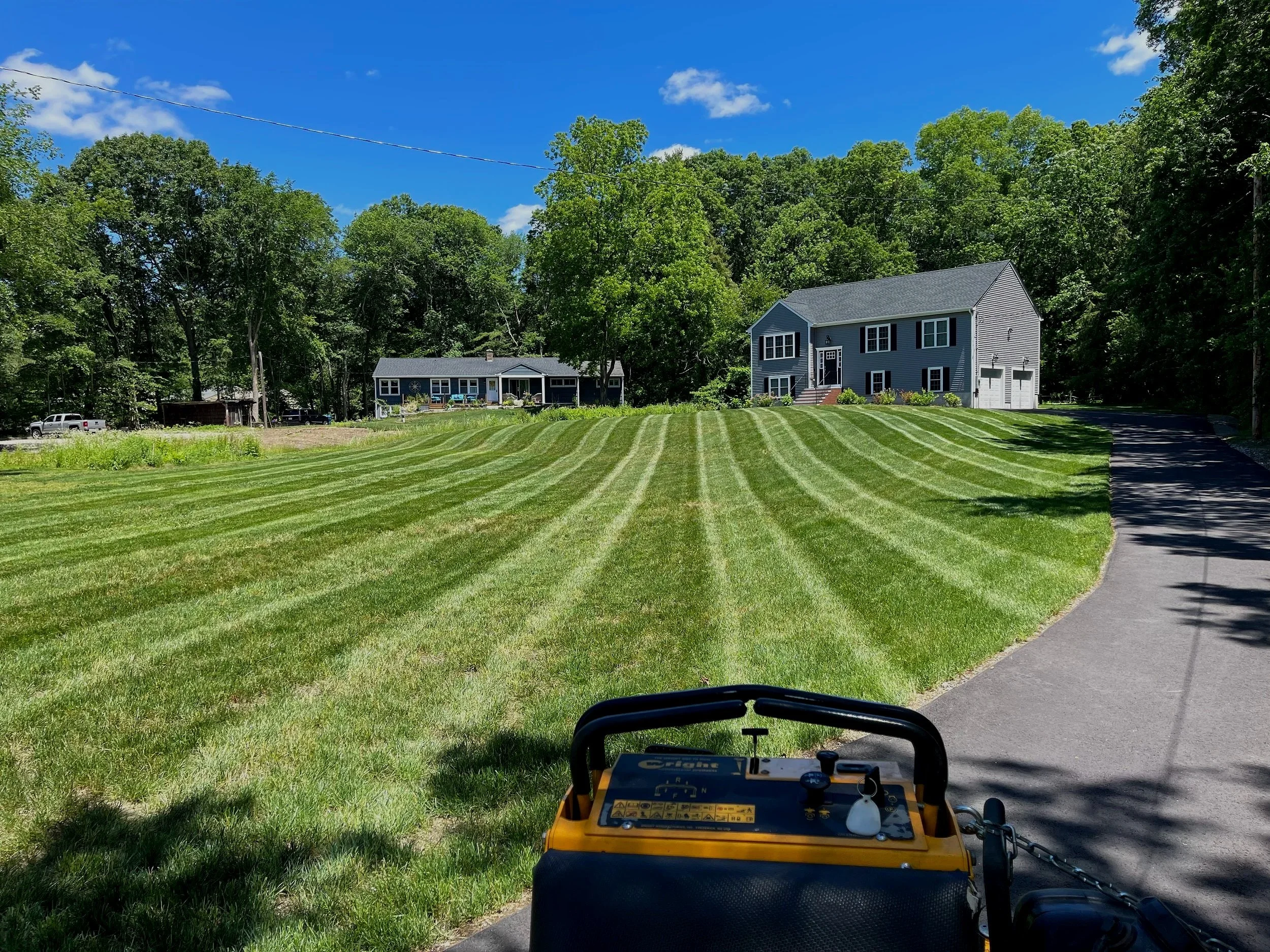 A suburban yard with freshly mowed striped grass, a house in the background, and a yellow lawn mower in the foreground. There is a driveway to the right and trees surrounding the yard.