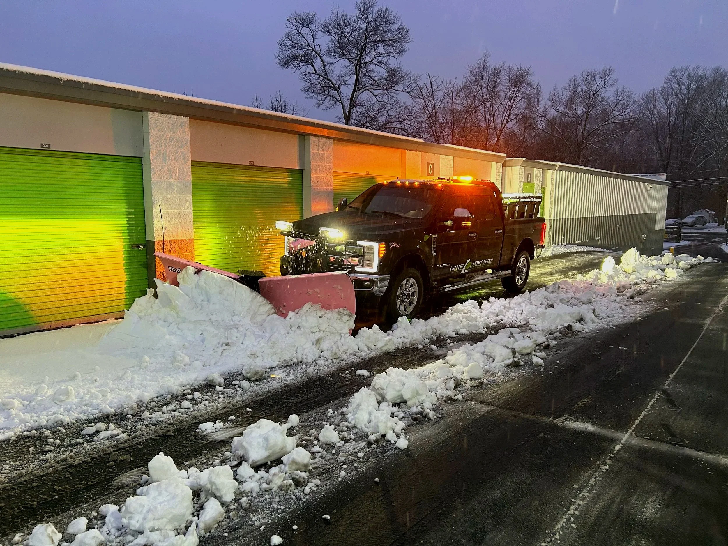 A black pickup truck with a snowplow attached, clearing snow from a parking lot at dusk. Snow is piled along the sides of the cleared area, and there are storage units with green doors in the background. Bare trees and a partly cloudy sky are visible