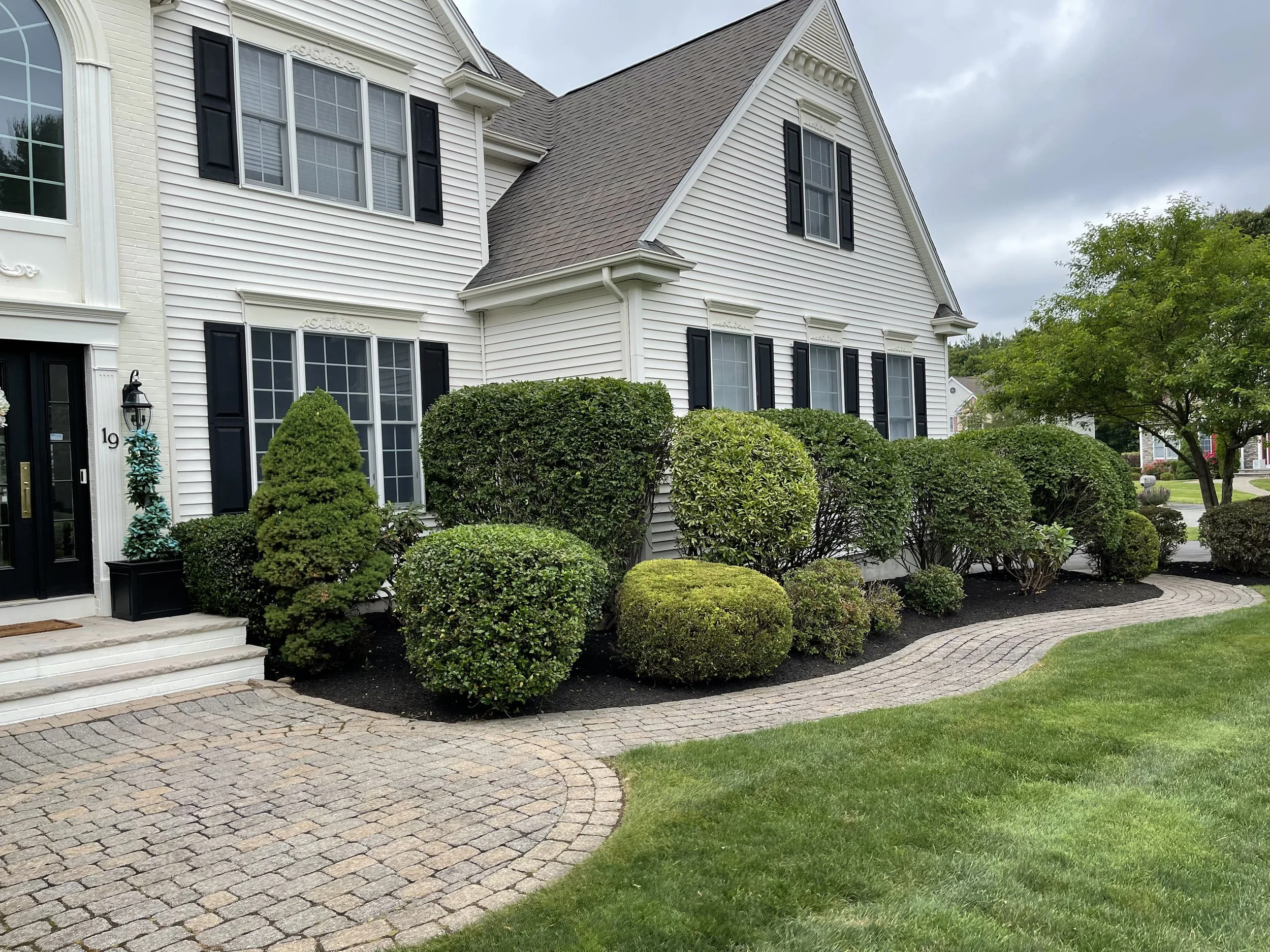 Front yard of a white two-story house with black shutters, surrounded by manicured bushes and trees, with a brick walkway and lawn.