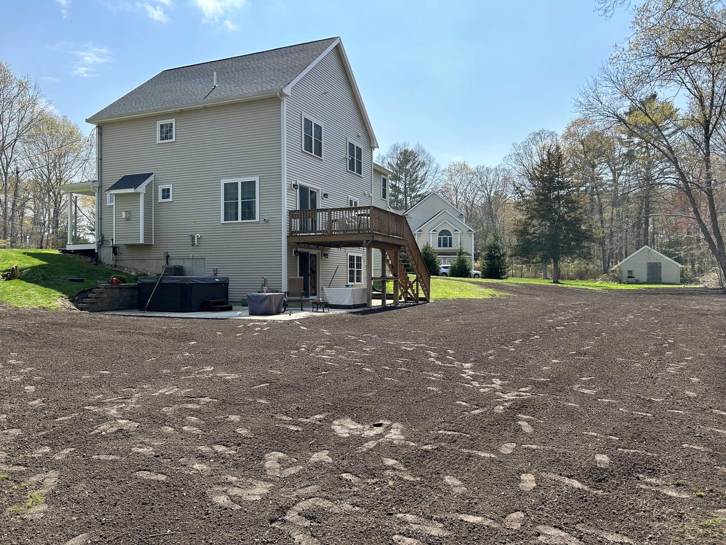 Backyard of a two-story house with a wooden deck, hot tub, and outdoor furniture, surrounded by trees and a large dirt area.