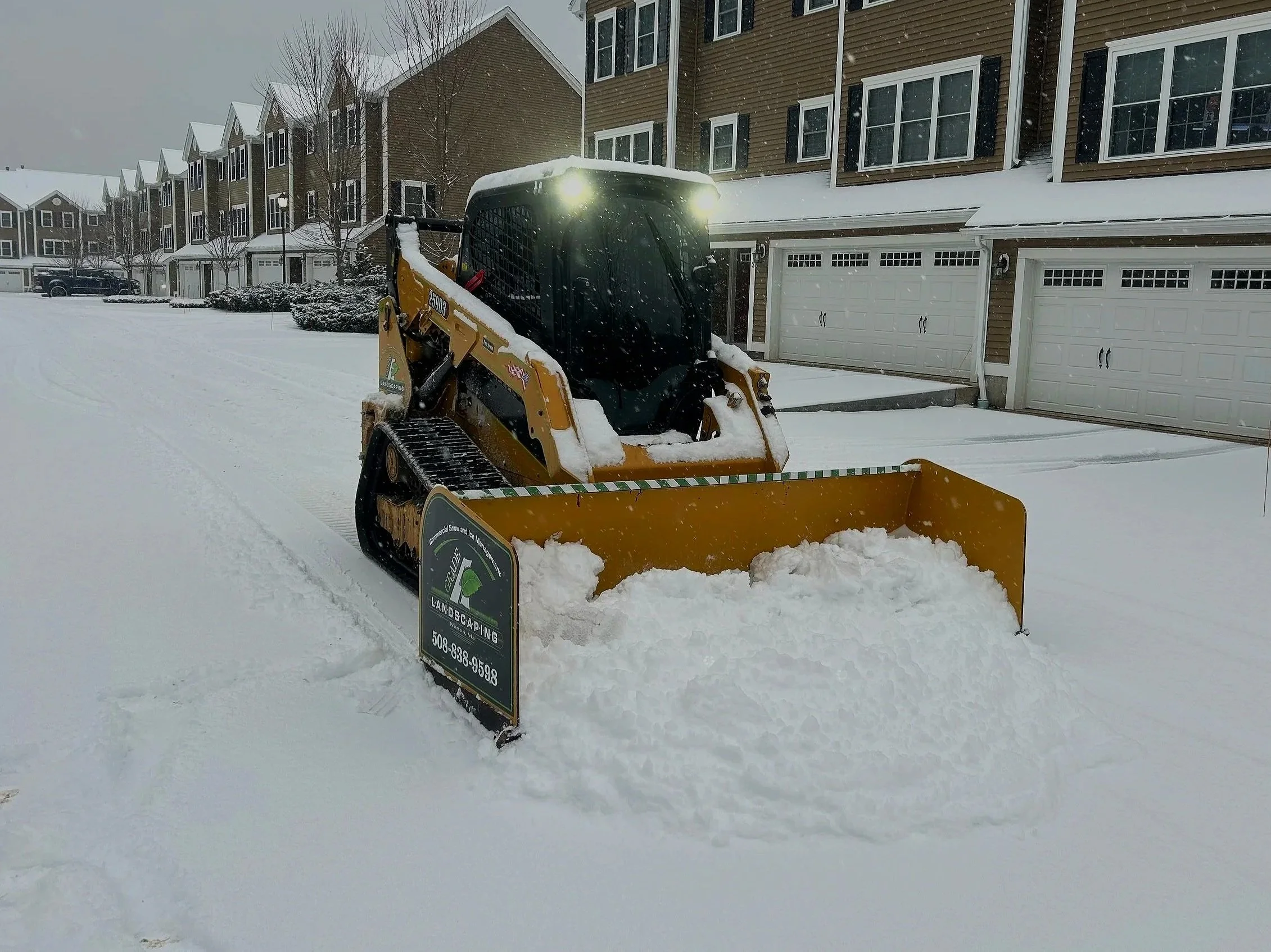 A yellow snowplow clearing snow from a residential street in a neighborhood, with modern houses and garages in the background.