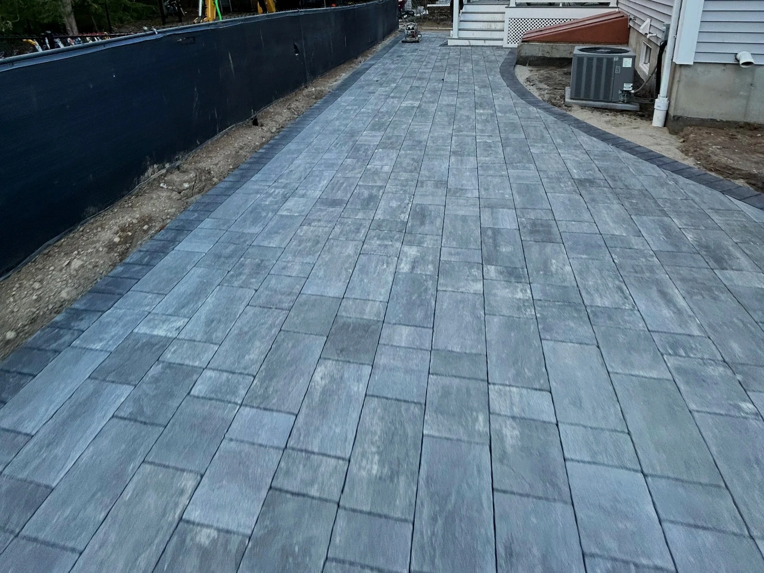 Newly paved gray stone walkway alongside a house, with construction materials and dirt on either side.