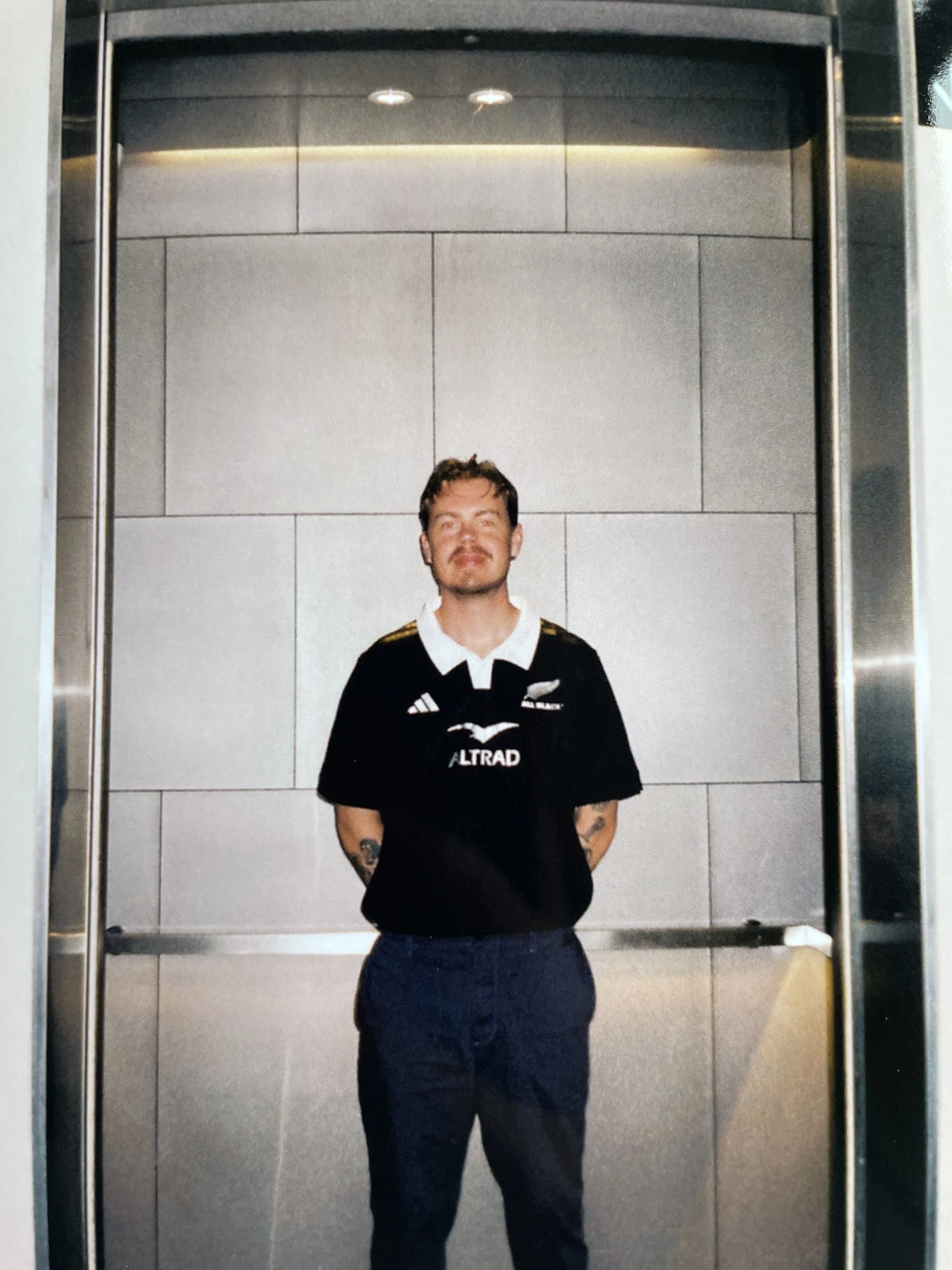A man with tattoos wearing a black sports jersey standing inside an elevator with gray tiled walls.
