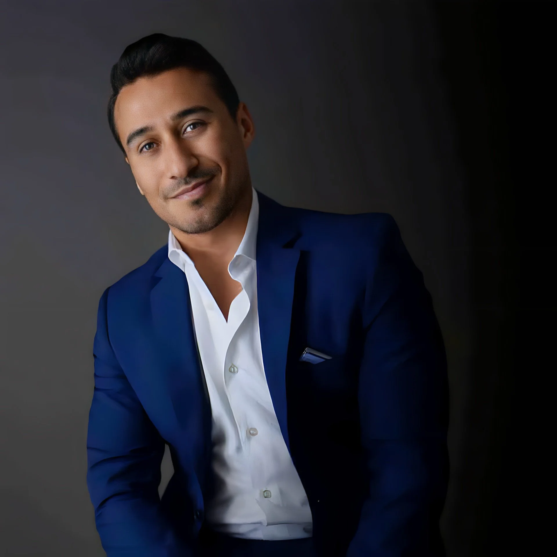 A young man with dark hair styled back, dressed in a navy blue suit and white shirt, smiling and looking at the camera against a dark background.
