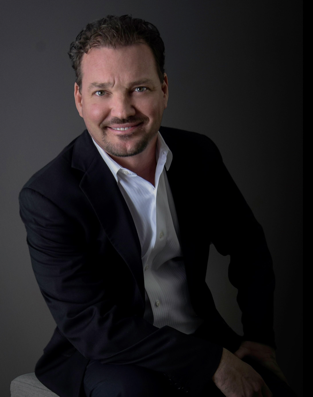 A portrait of a man with short dark hair and a beard, wearing a black blazer and white shirt, smiling at the camera against a dark background.
