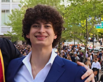 A young man with curly dark hair smiling outdoors in a crowd, wearing a white shirt and blue blazer.
